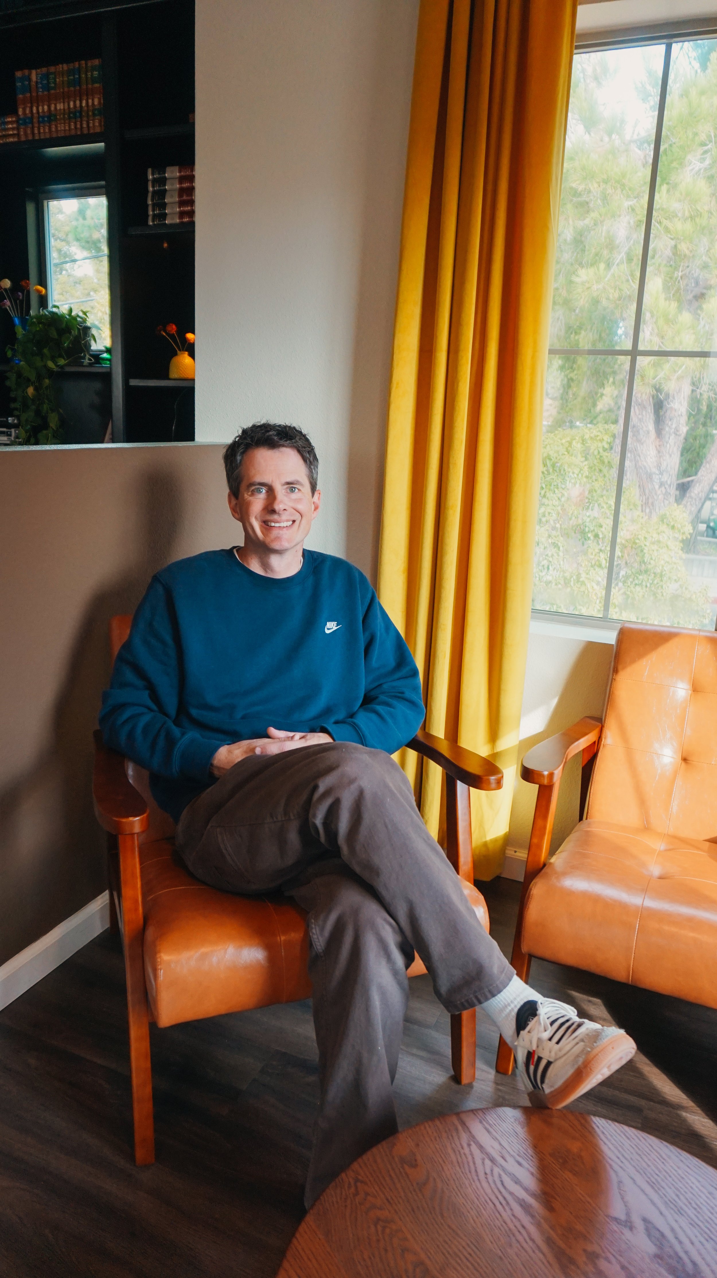 A man with short dark hair wearing a blue Nike sweatshirt, gray pants, and white sneakers, sitting on an orange leather chair near a window with yellow curtains, in a room with dark wood flooring and a black bookshelf. Outside the window, trees are visible.
