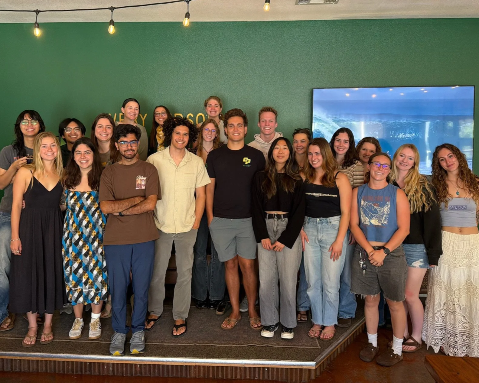 A group of young adults, both men and women, standing on a stage, smiling for a photo. They are in a room with green walls and a large screen on one side showing a scenic background. The group is casually dressed, with some wearing glasses and others with distinctive hairstyles.