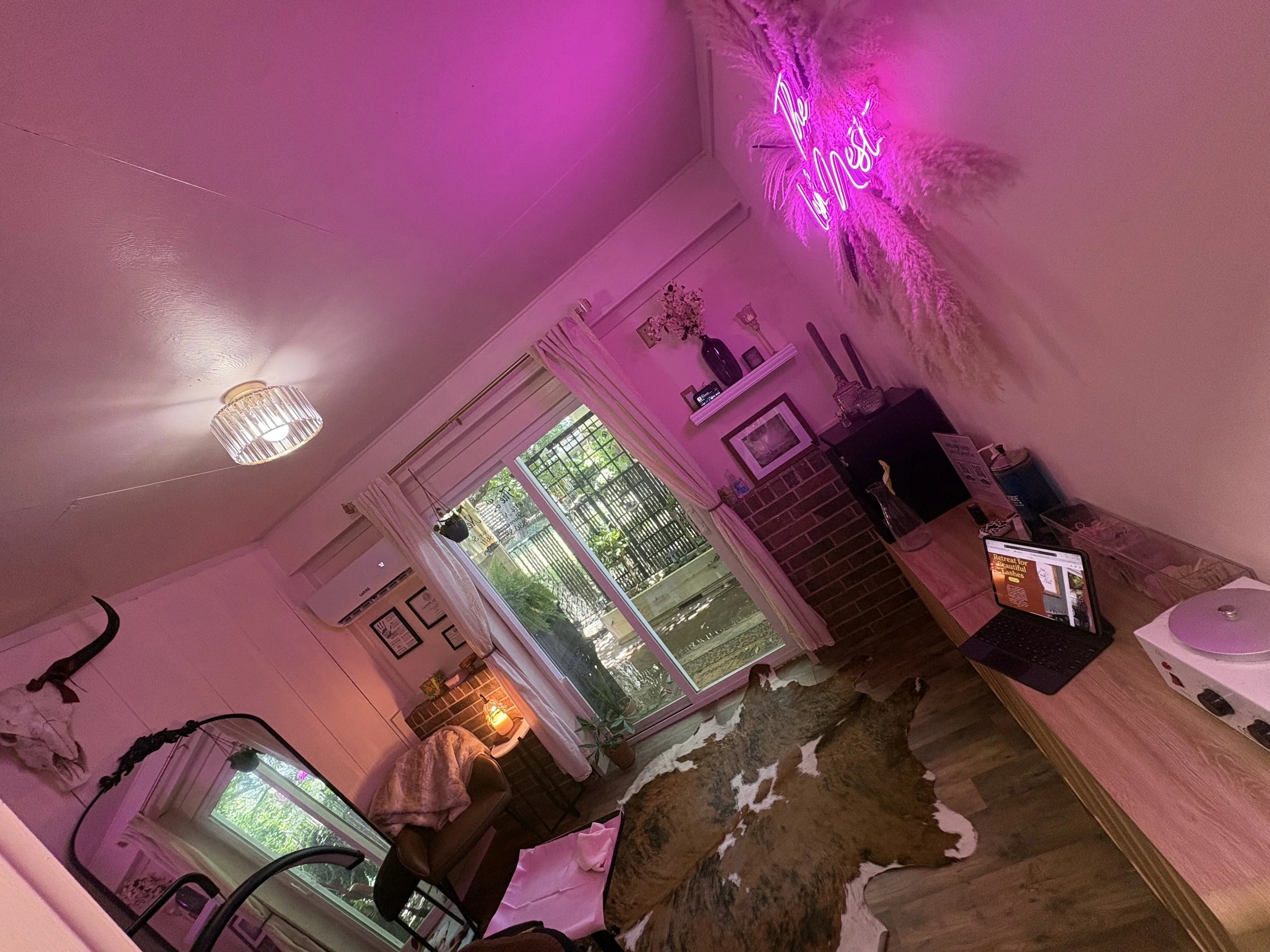 A cozy living room decorated with pink neon lights spelling out 'Hello' on the wall, with a potted plant, shelves with framed pictures, and a window with curtains. There is a desk with a laptop and a cowhide rug on the floor.
