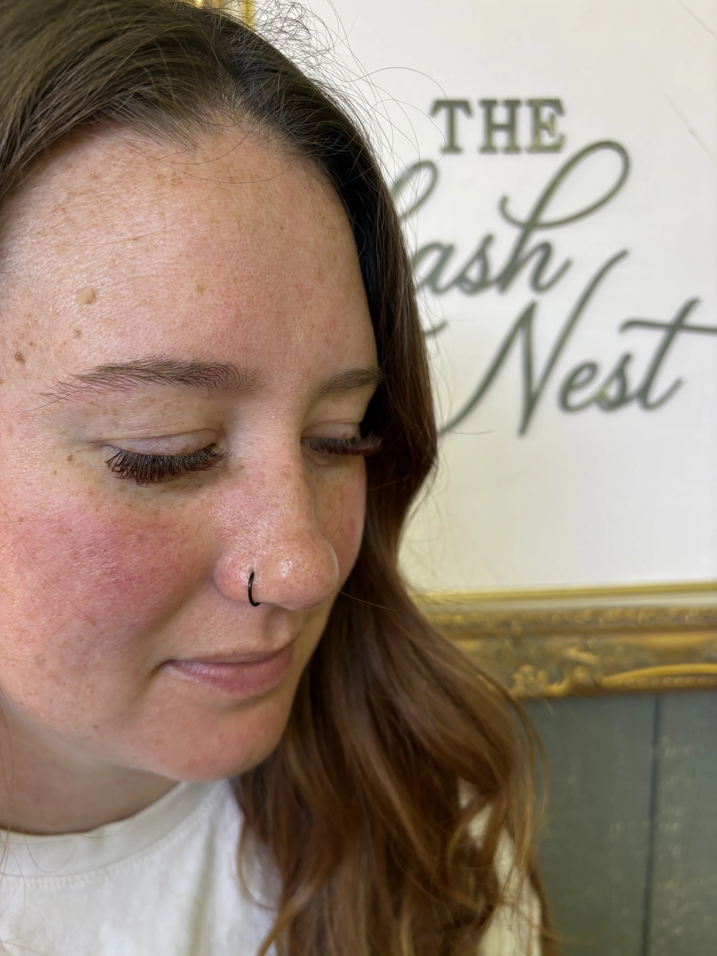 Close-up of a woman's face showing her forehead, eyebrow, closed eyes, and part of her nose with a nose ring. She has red hair and some freckles and moles on her skin. The background features a wall with a sign that reads 'THE BRUSH N' at the top.