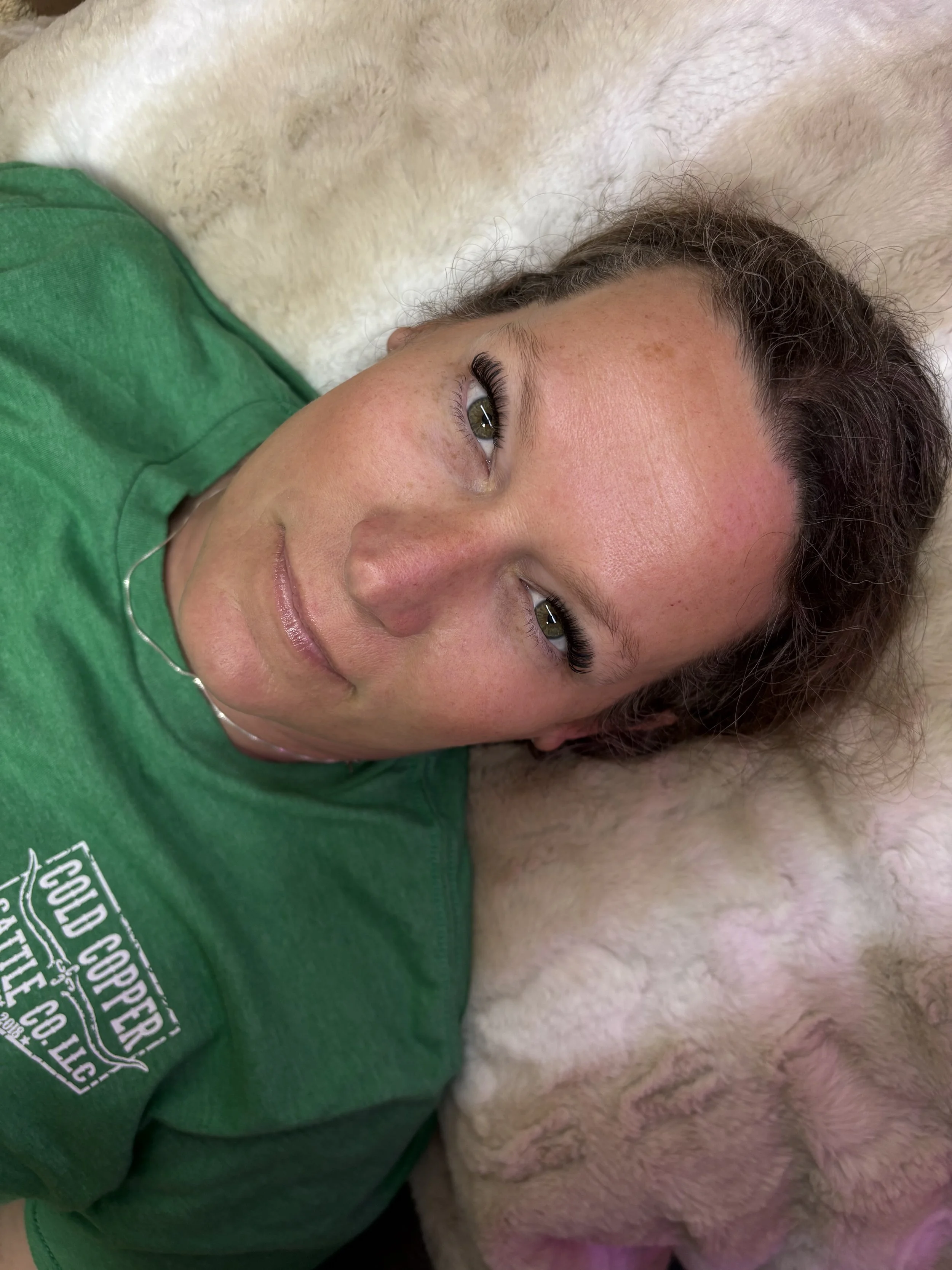A woman lying down on a soft surface next to a large dog, with her head resting on the dog's body. She is wearing a green t-shirt and has dark hair.