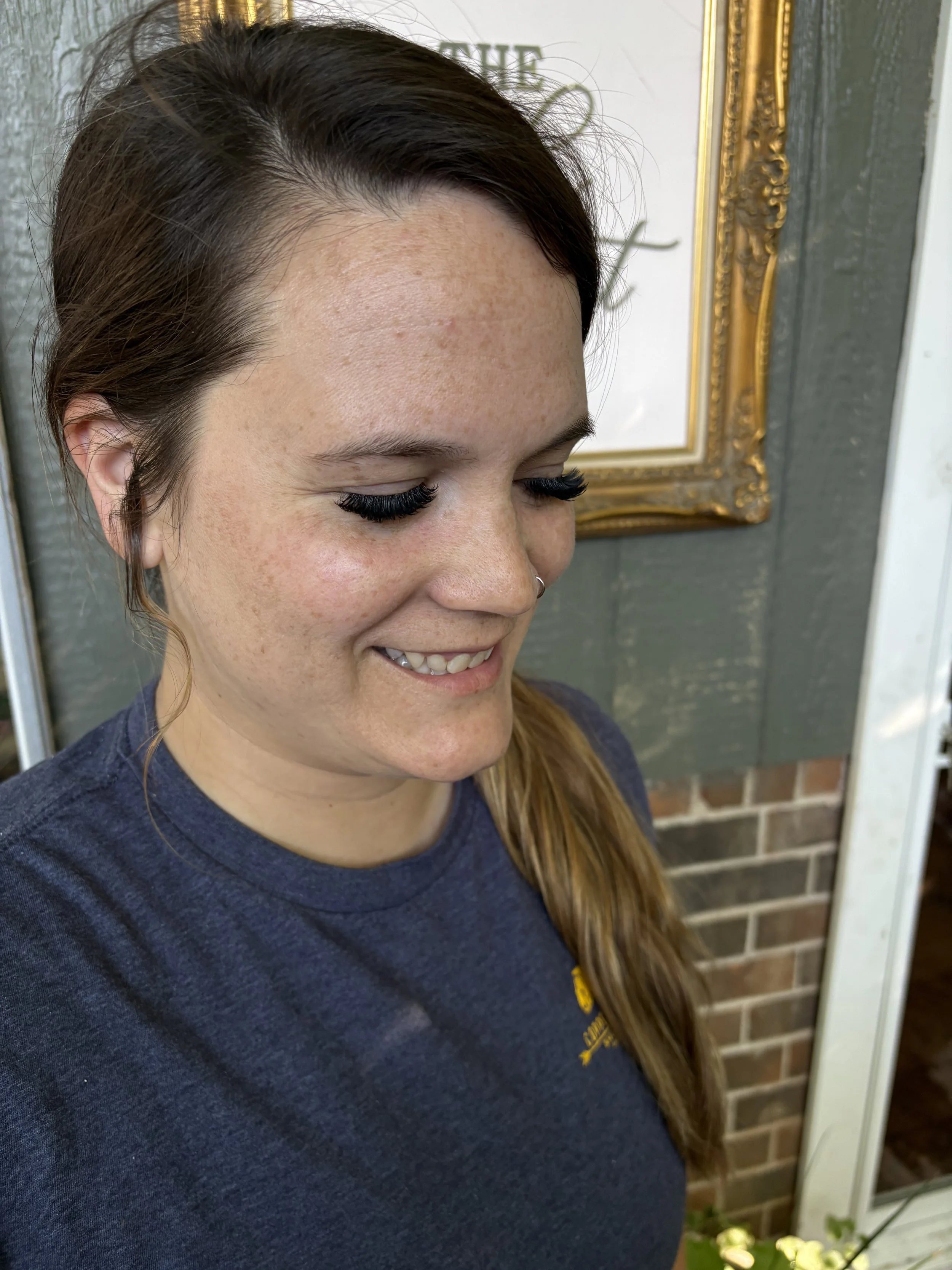 Close-up photo of a smiling woman with long red hair, dark eyelashes, and freckles, standing in front of a framed mirror and a brick wall, wearing a navy blue shirt.