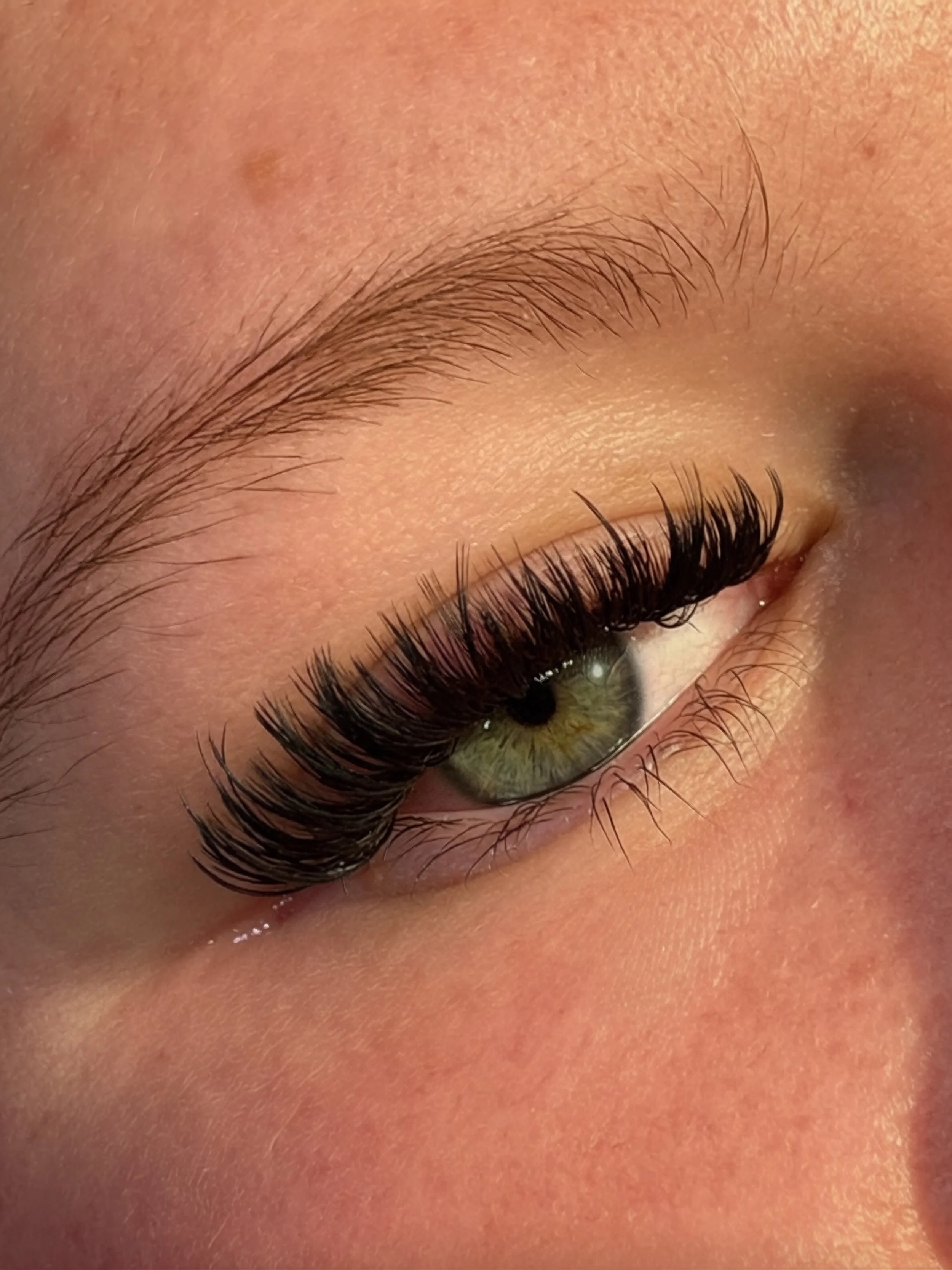 Close-up of a person's eye with long, curled eyelashes and a green iris, surrounded by smooth skin.