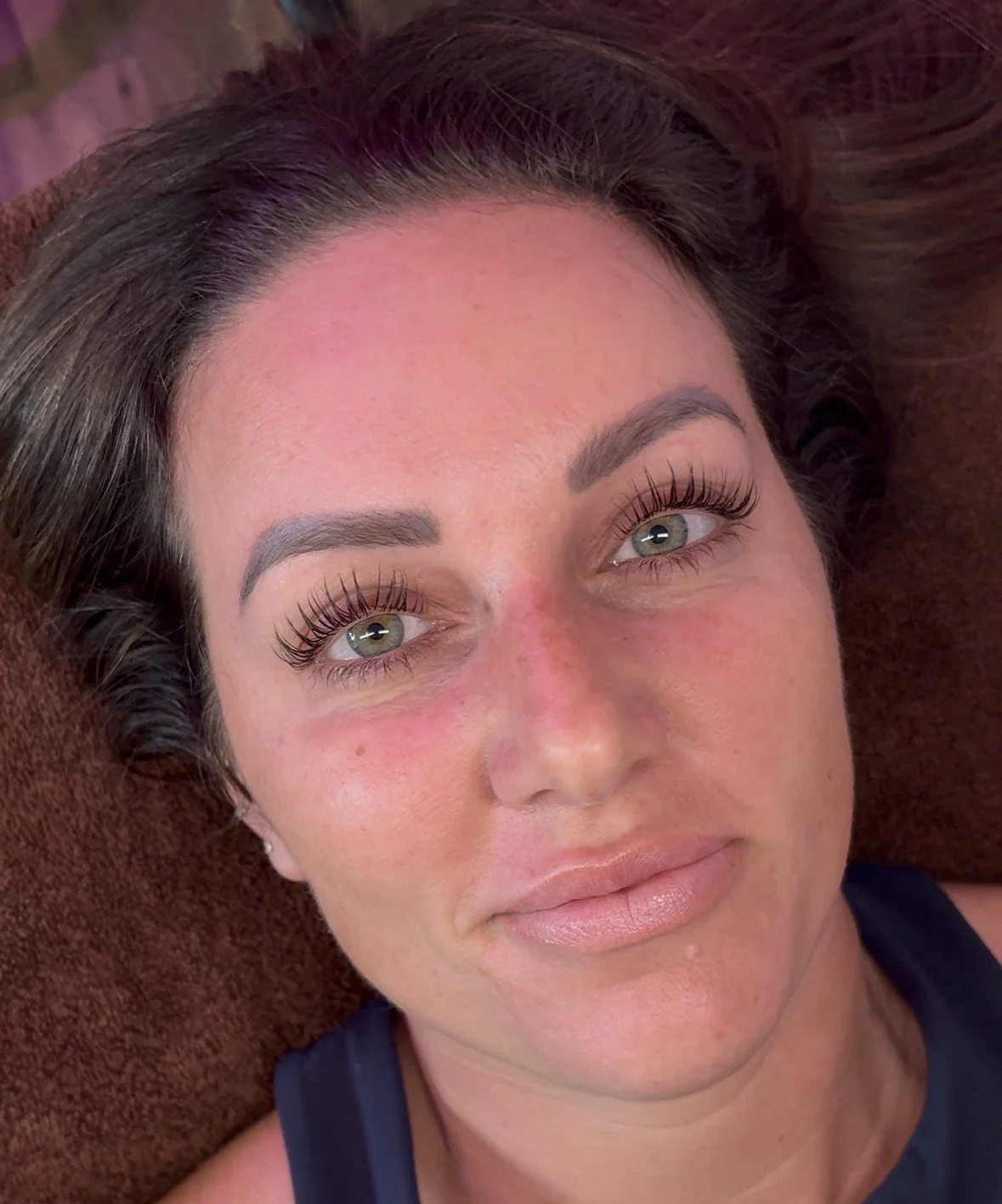 Close-up of a woman lying down on a brown surface, with gray-green eyes, long eyelashes, bold eyebrows, and light makeup, smiling softly.