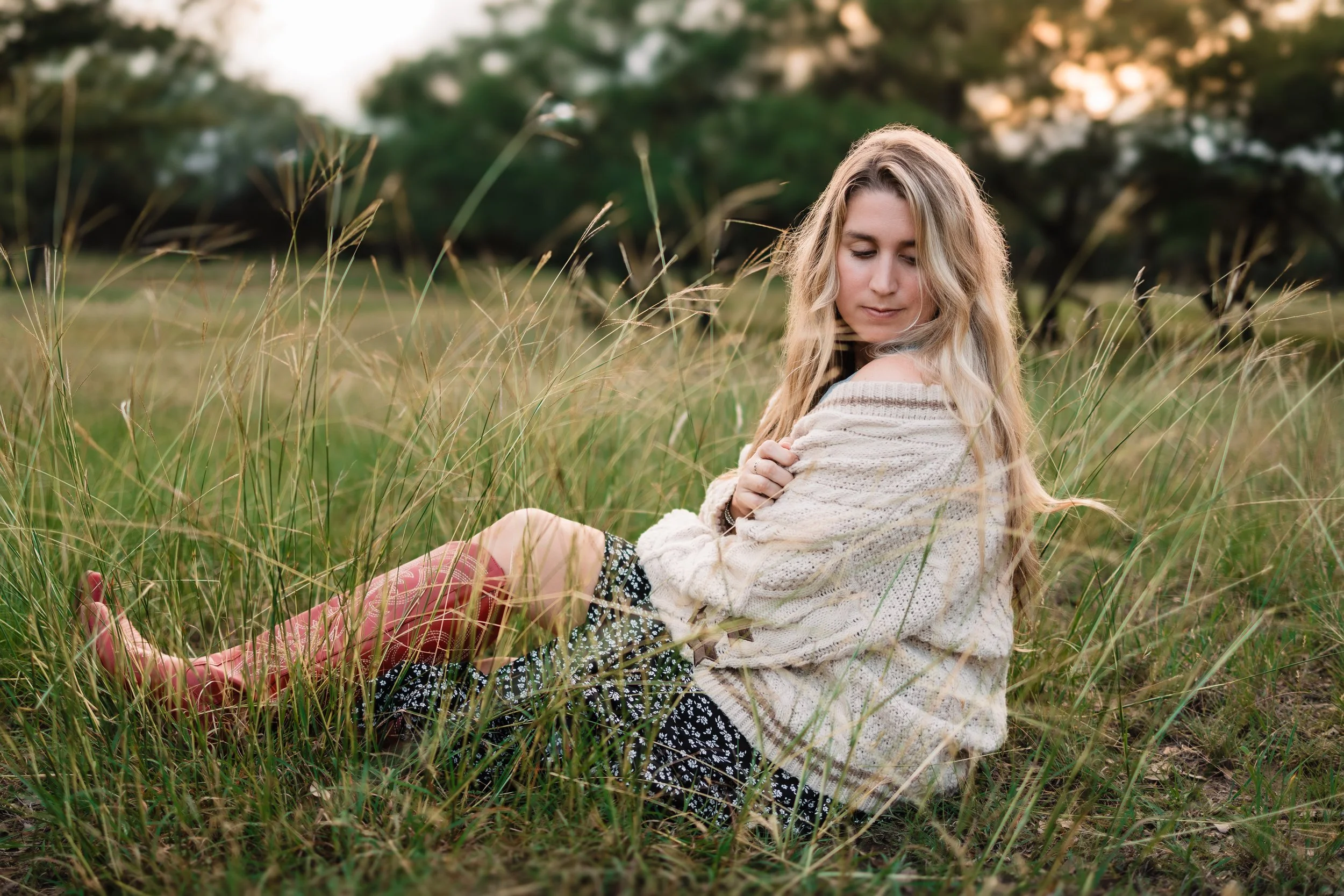 A young woman with long blonde hair sitting in tall grass in a field during sunset, wearing a cream sweater and patterned skirt.