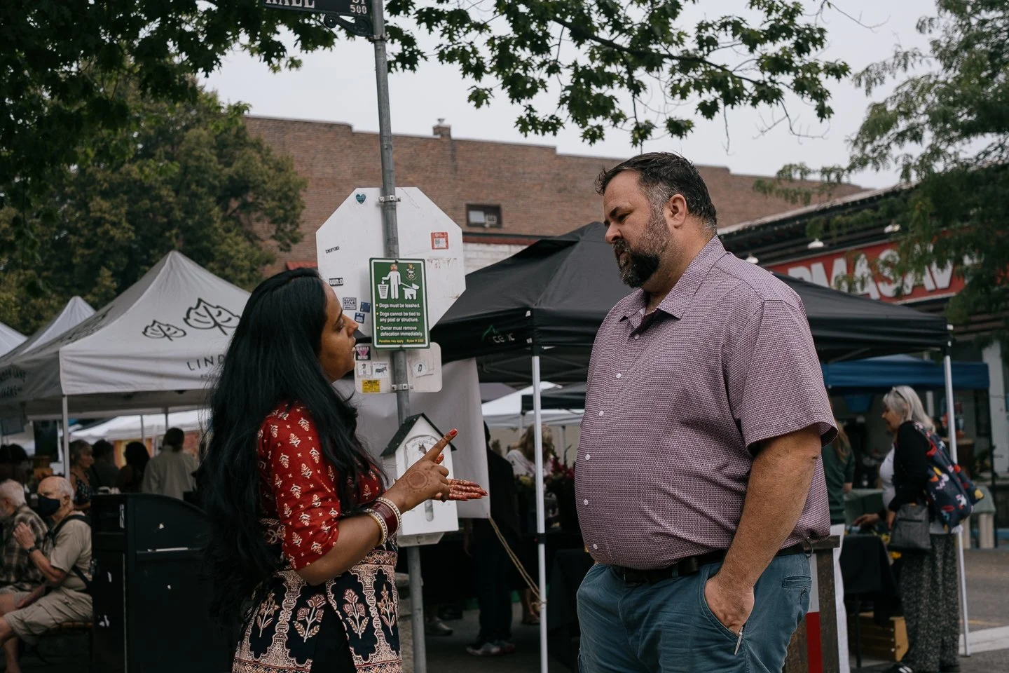 A woman with long black hair and a red blouse is talking to a tall man with a beard and short hair, wearing a purple short-sleeve shirt, at an outdoor event with tents and people in the background.