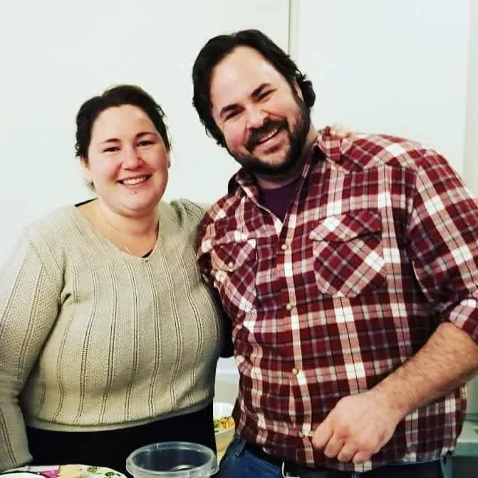 A smiling woman and man standing together indoors, with the woman wearing a beige sweater and the man in a red plaid shirt.