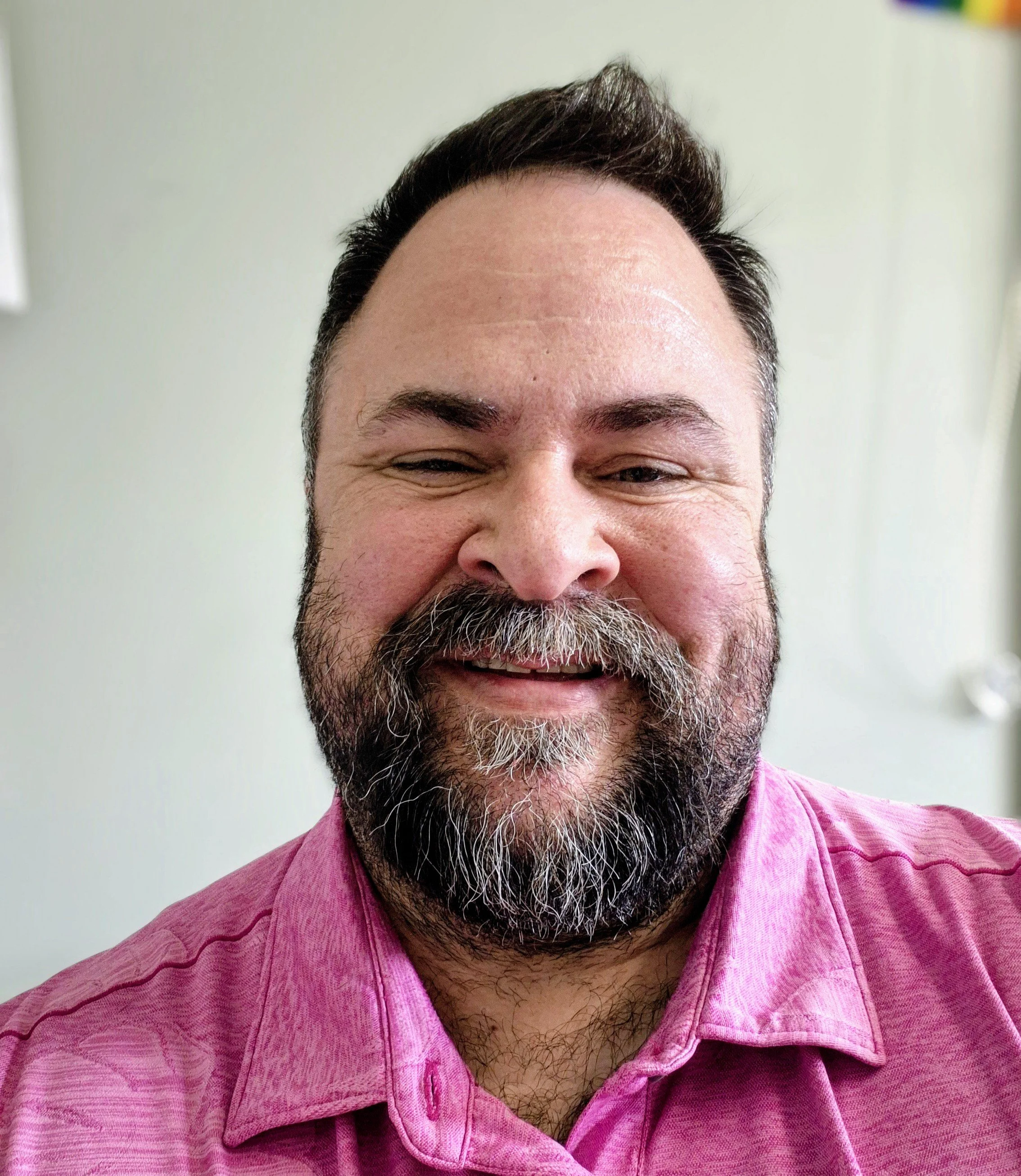 Close-up of a smiling man with a beard and mustache, wearing a pink button-up shirt, against a plain light-colored background.