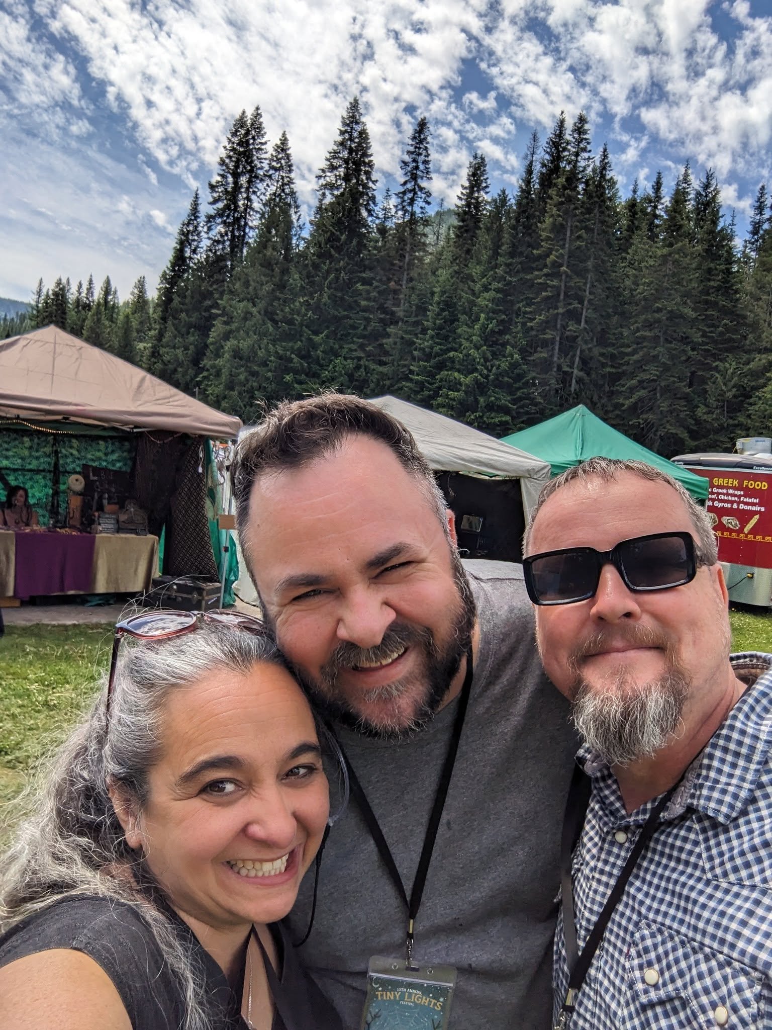 Four people smiling and taking a selfie outdoors at a festival with trees, tents, and food stalls in the background.