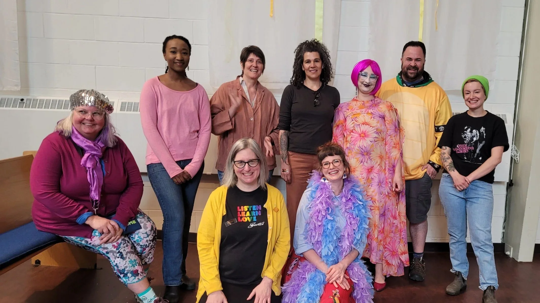 Group of eleven diverse people gathered indoors, some dressed in colorful and playful costumes, smiling for a photo.