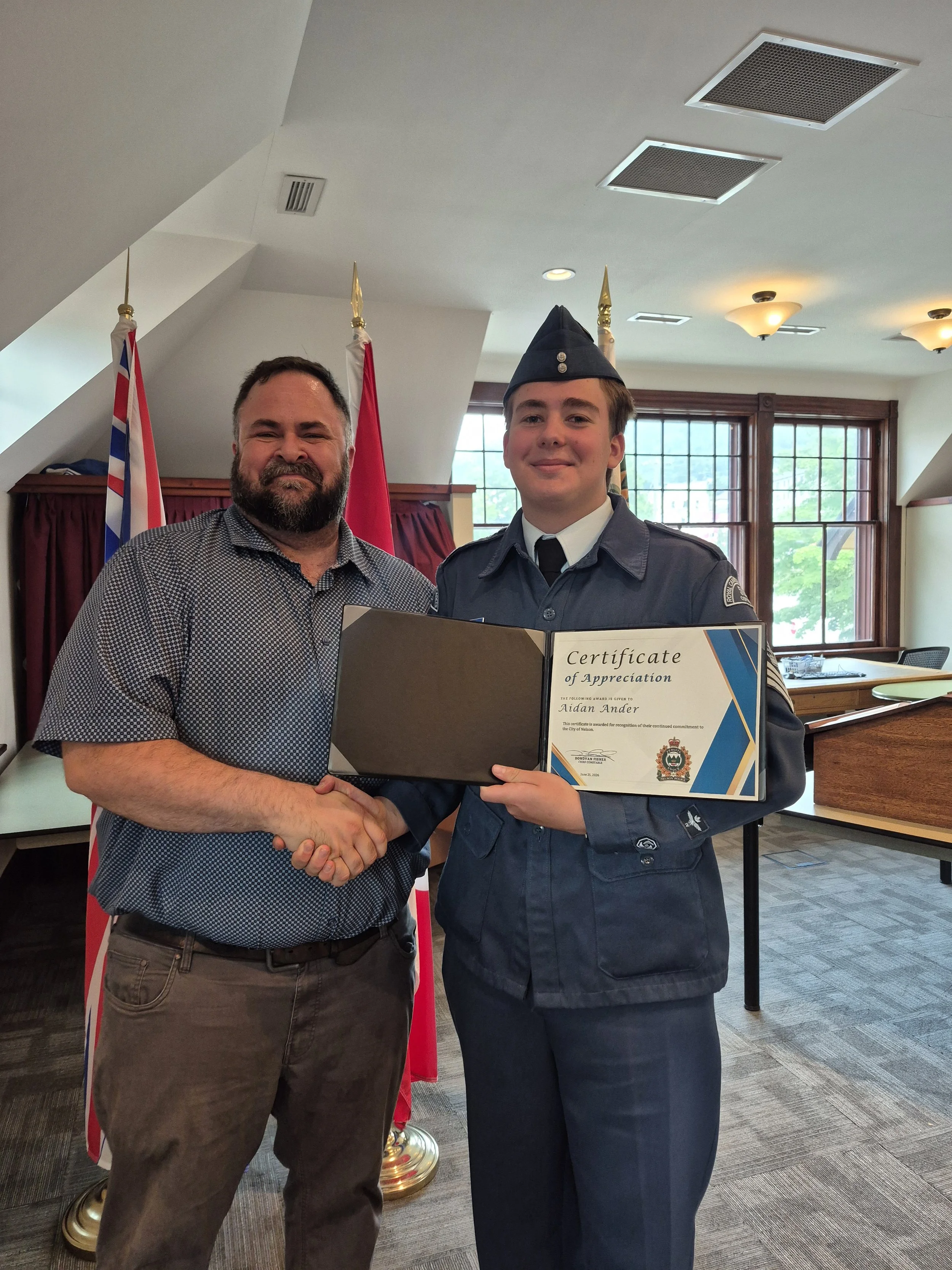A man and a young man in a police uniform shaking hands indoors, holding a certificate of appreciation. Behind them are flags and large windows with natural light.