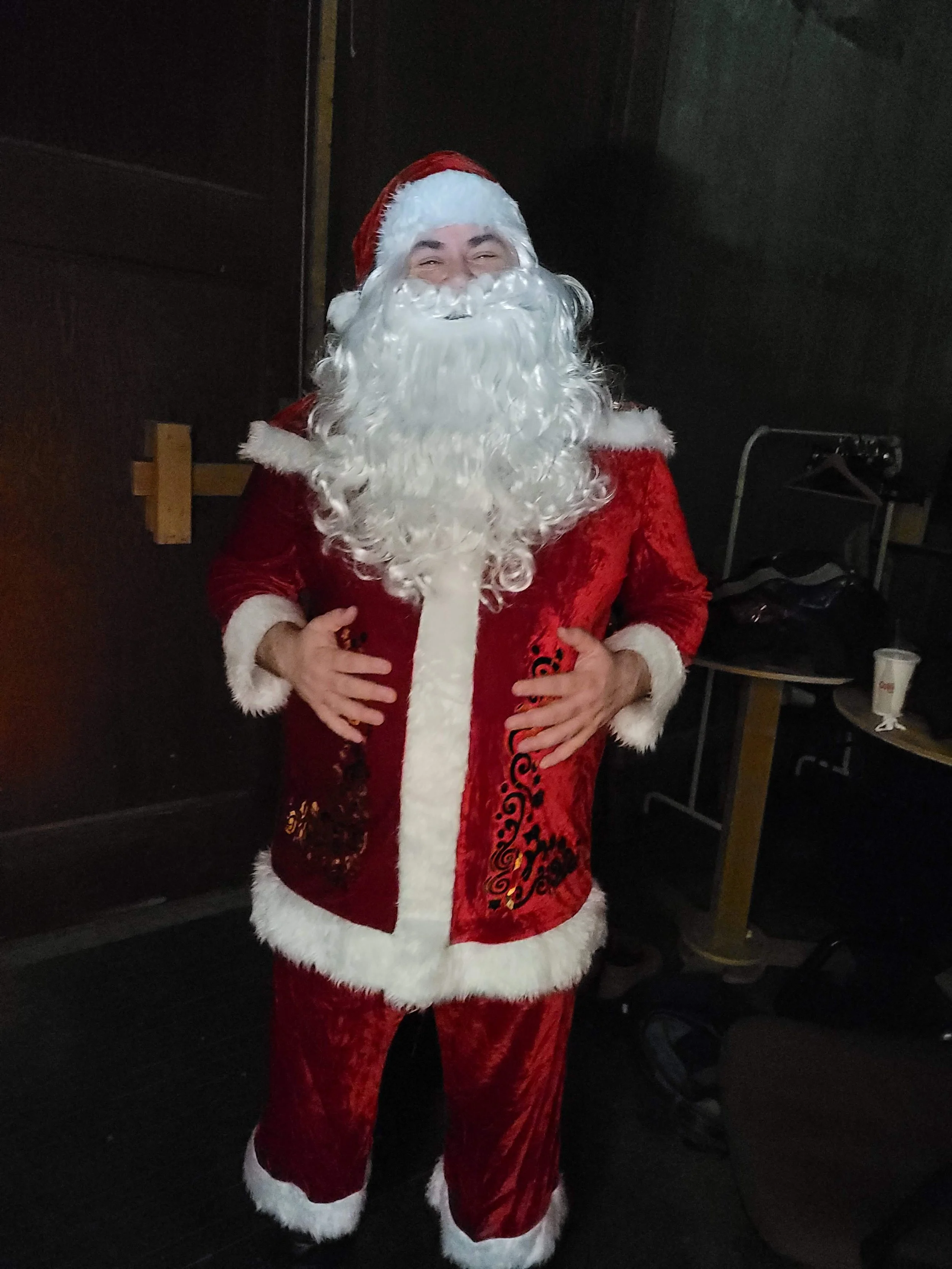 A person dressed as Santa Claus standing indoors in front of a black wall, wearing a red velvet suit with white fur trim, a matching red hat with white fur, and a large white beard.