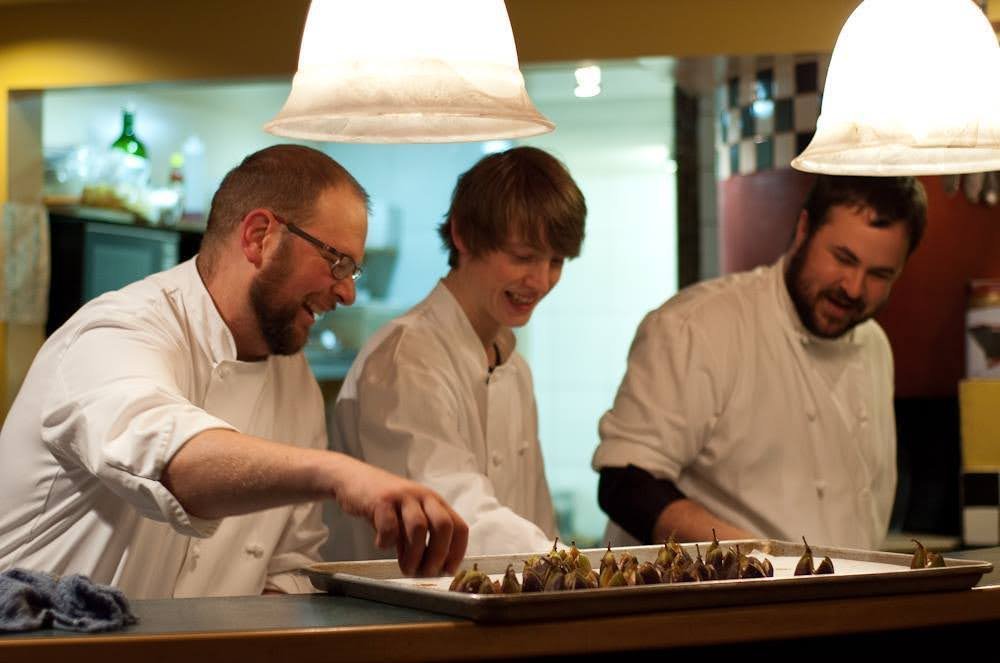 Three chefs in a restaurant kitchen preparing a tray of appetizers, smiling and working together.