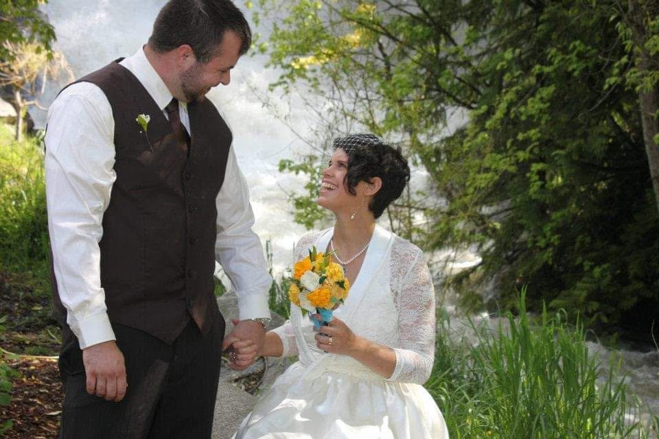 A bride and groom holding hands outdoors near a stream, smiling at each other. The bride wears a white lace dress and a birdcage veil, holding a bouquet of yellow and white flowers. The groom wears a white shirt and dark vest, with a boutonniere on h