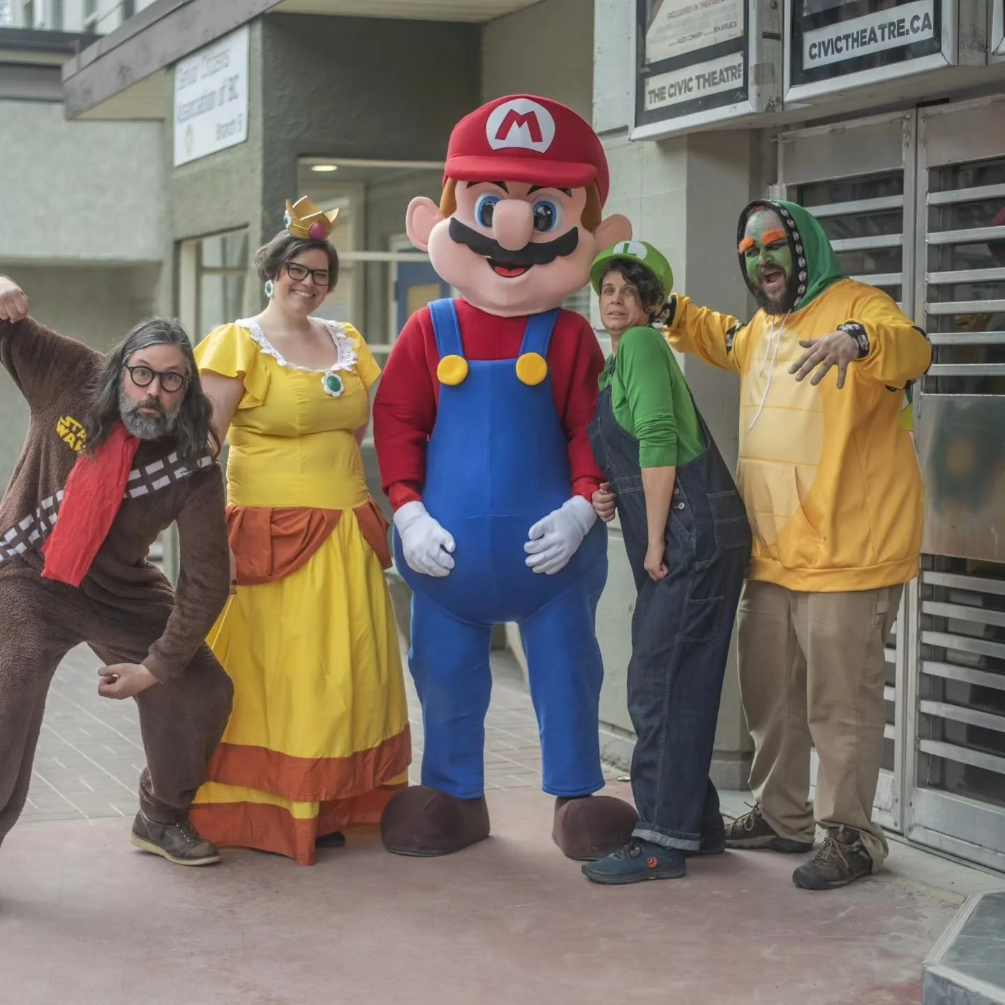Group of people in costume, including a person dressed as Mario from video games, posing together outside a building with signs and a metallic door.
