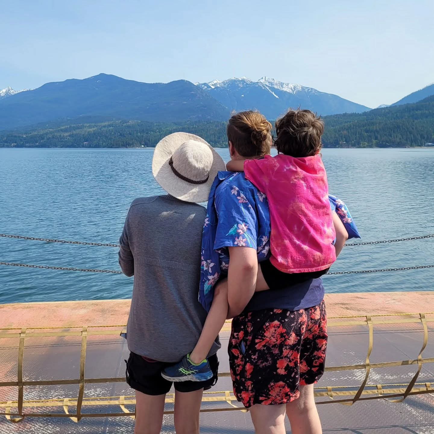 Family looking at a lake with mountains in the background, woman holding a child on her back, man wearing a sun hat and sweater, scenic mountain landscape with snow on peaks.
