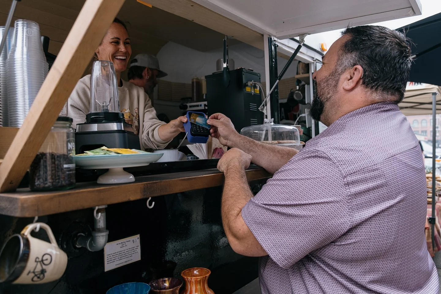 A man paying with a credit card to a woman working at a food stand, smiling, in an outdoor market setting.