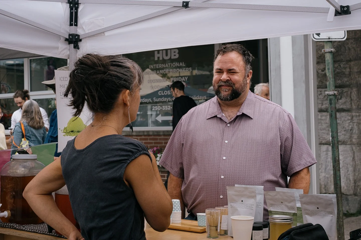 A woman and a man are conversing at a small outdoor marketplace stall with various jars and packages, under a white canopy tent.