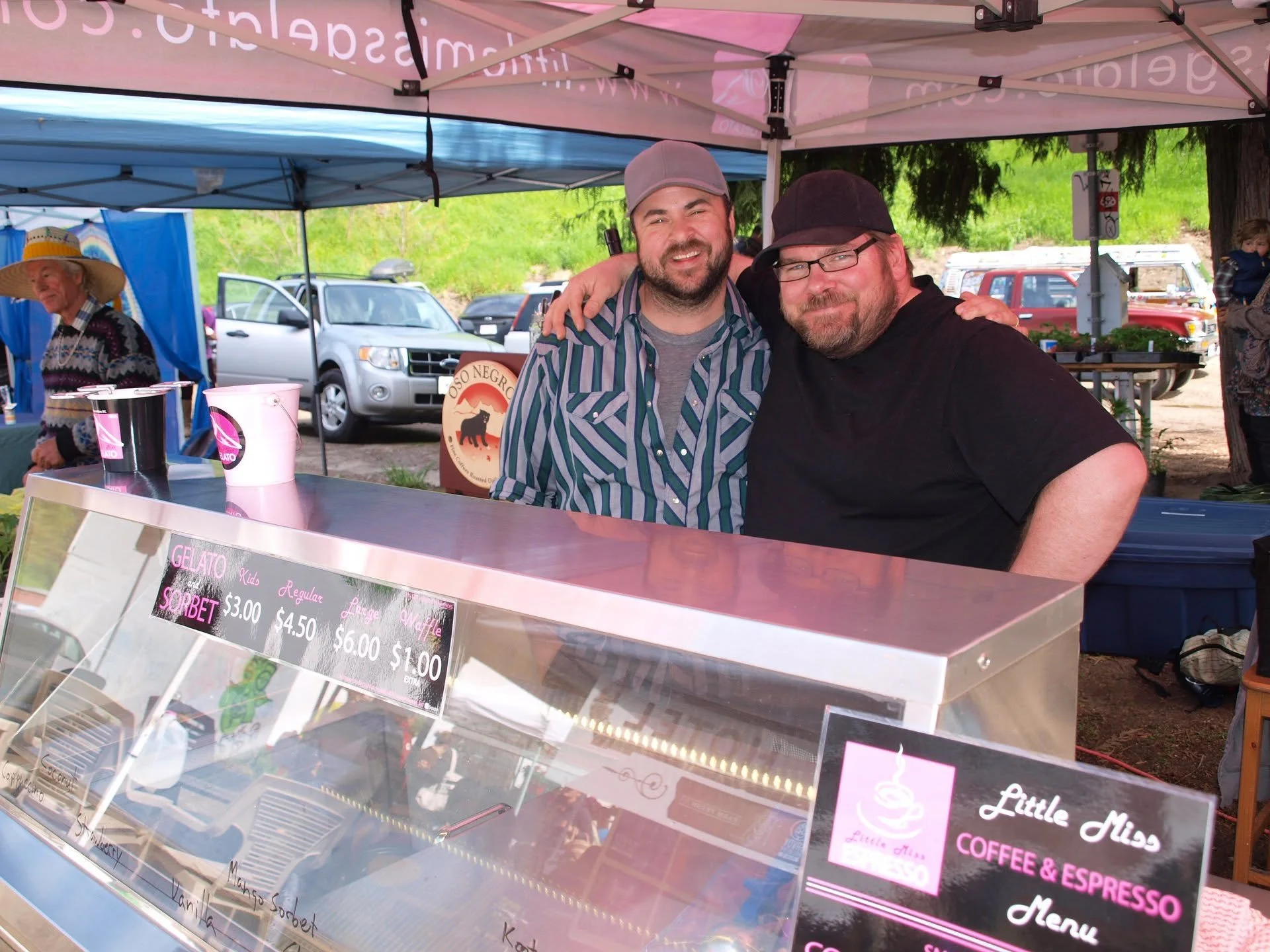 Two men smiling and posing for a photo at an outdoor market stall under a pink and blue canopy, with cars parked in the background, and a woman wearing a large straw hat standing nearby.