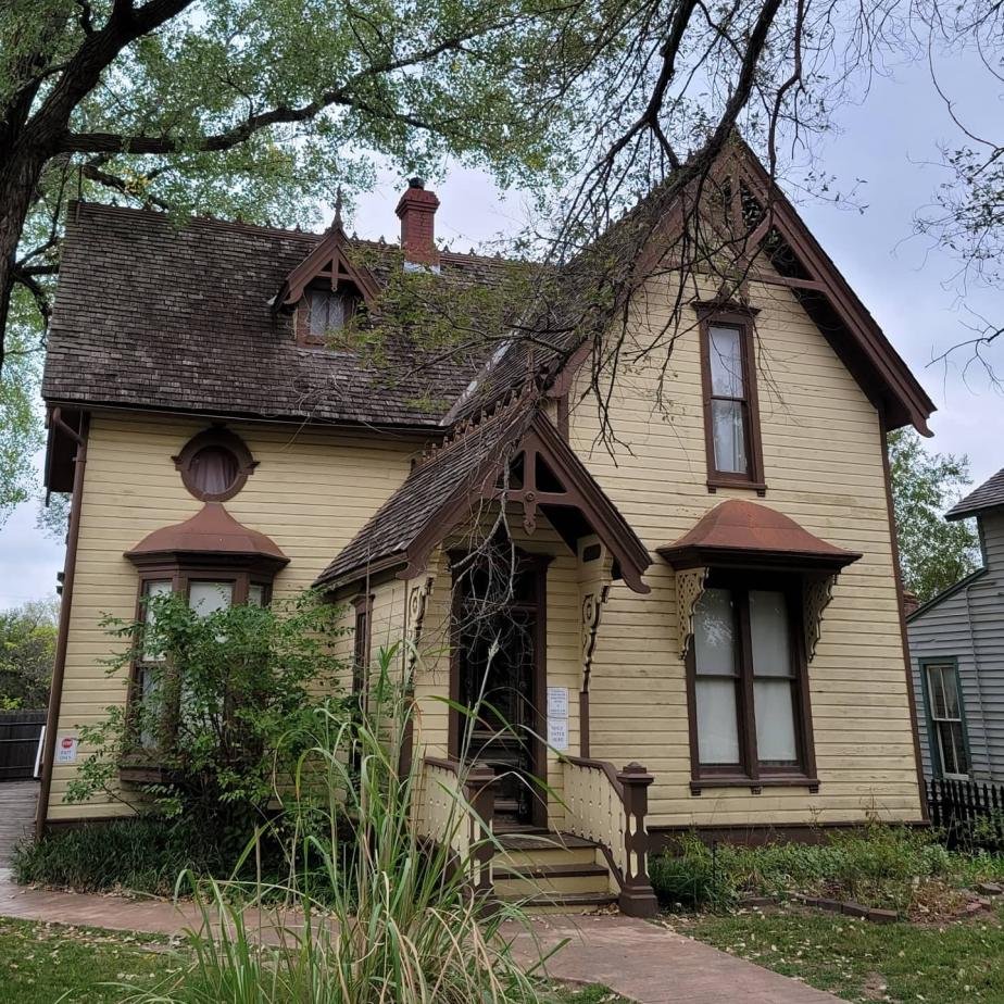 Menger House in Wichita, Haunted location. A two-story Victorian-style house with beige siding, decorative brown trim, and a steep roof, surrounded by trees and bushes.