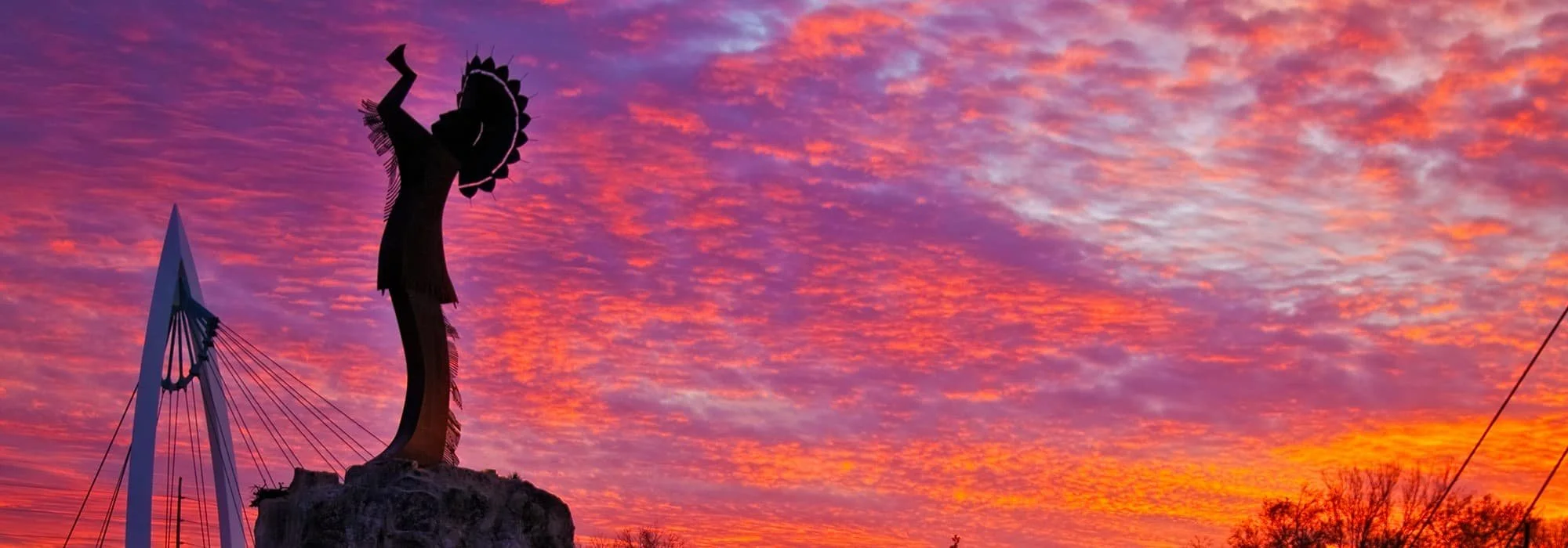 Keeper of the plains in Wichita at Sunset.  Silhouette of a sculpture against a vibrant sunset sky with pink, purple, and orange clouds. A modern bridge is visible to the left, and trees are silhouetted at the bottom.