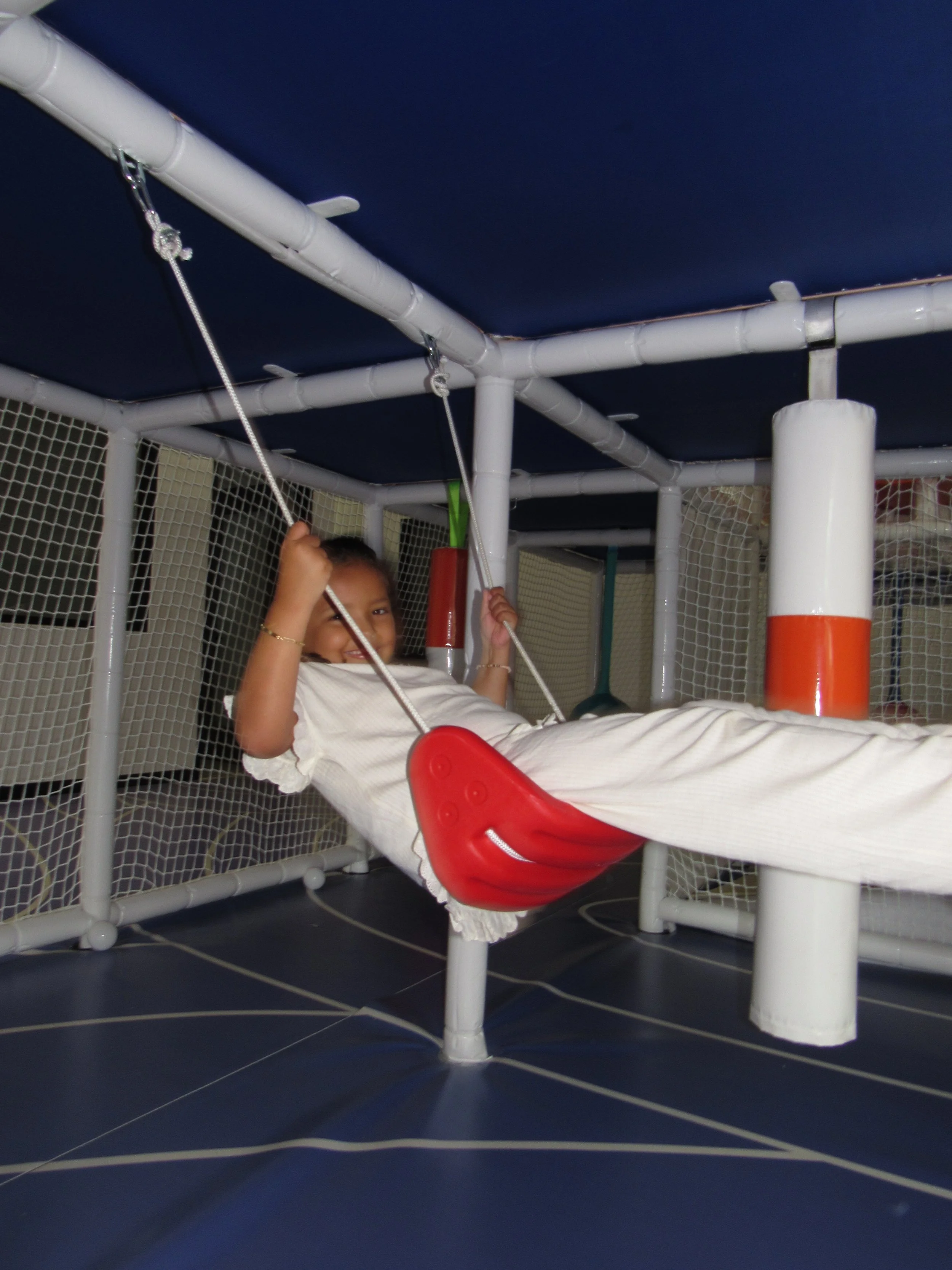 Child lying on a swing inside a play structure with white pipes, red seat, and netting walls, in an indoor play area with dark blue ceiling.