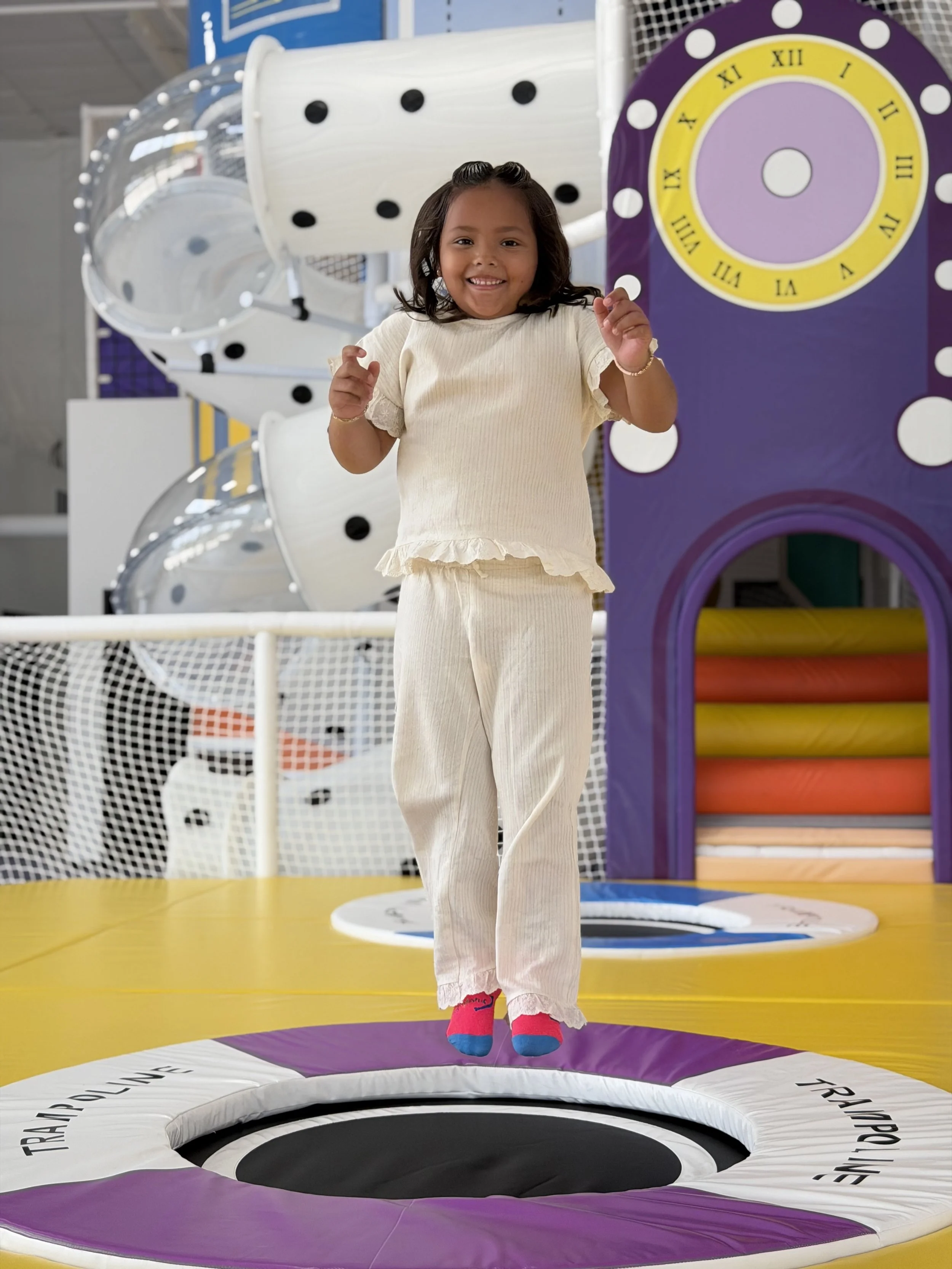 A young girl is jumping on a trampoline inside an indoor play area, smiling and holding a small object in her right hand. She is wearing a cream-colored dress with ruffled sleeves and pink socks with blue accents. In the background, there are colorful structures and safety nets.