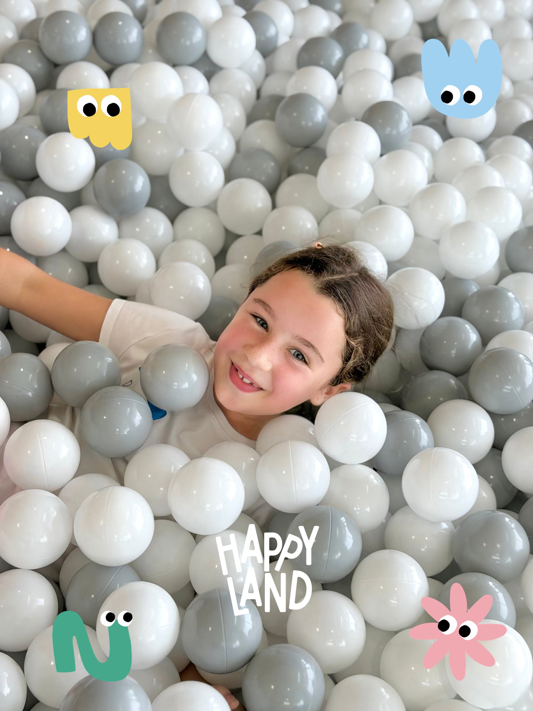 A girl lying in a ball pit filled with white and gray plastic balls, decorated with colorful cartoon stickers of a blue bunny, pink flower, green snake, and a yellow character. The girl is smiling and looking at the camera.