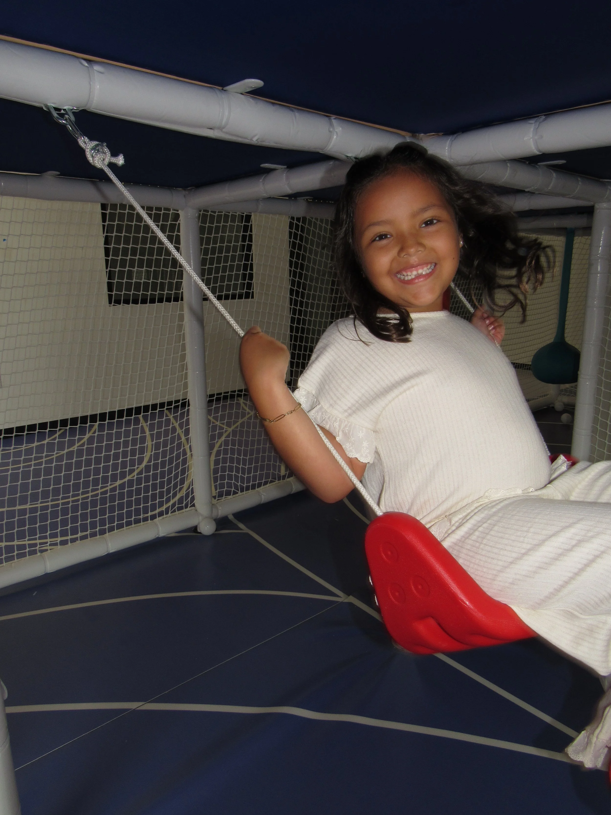 A smiling young girl wearing a white dress, sitting on a red swing attached to a play structure with netting. The play structure has a blue roof and white pipes.