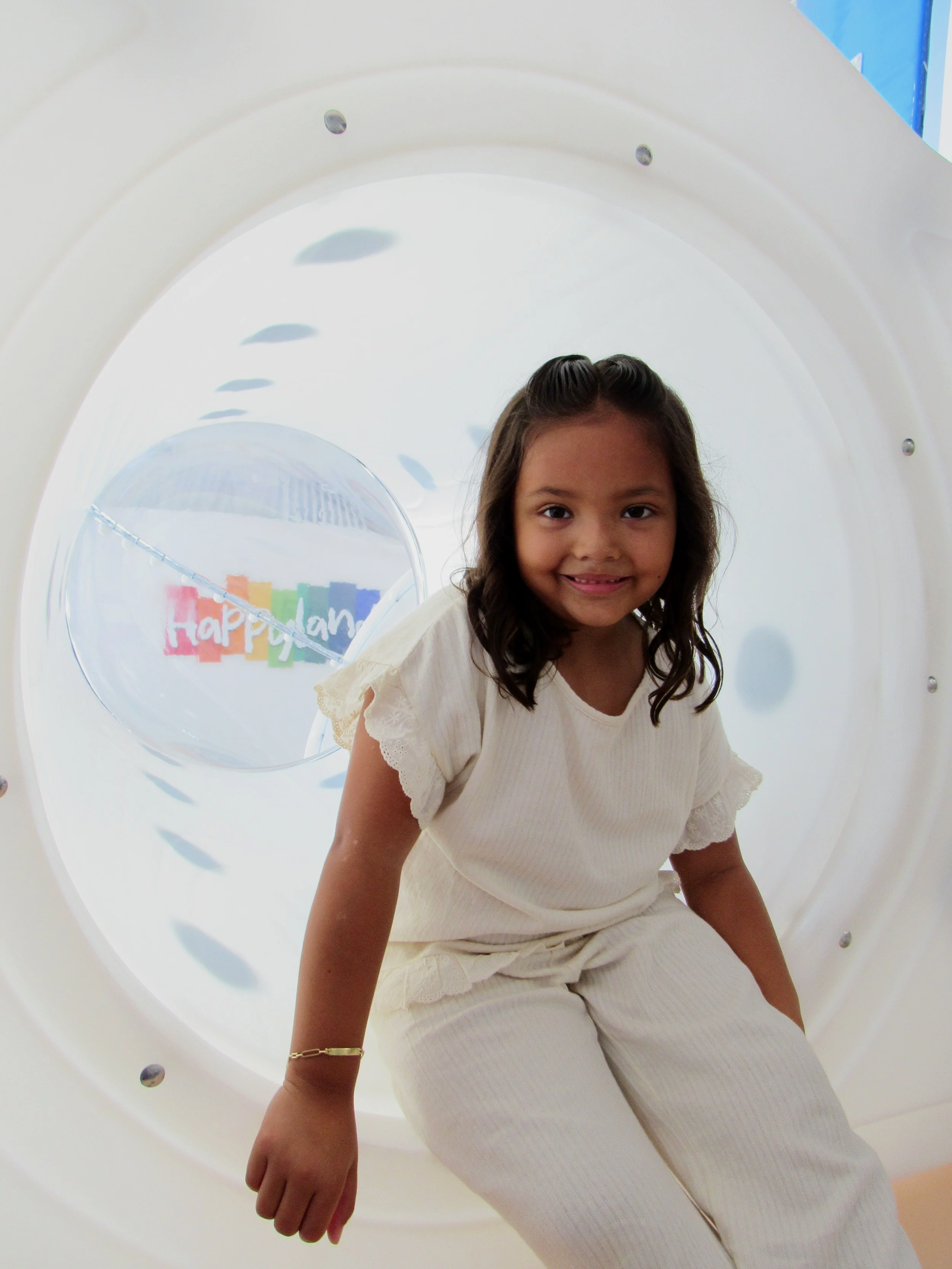 A young girl with long dark hair, wearing a cream-colored dress with lace details, sitting inside a white circular tunnel with a smiling expression.