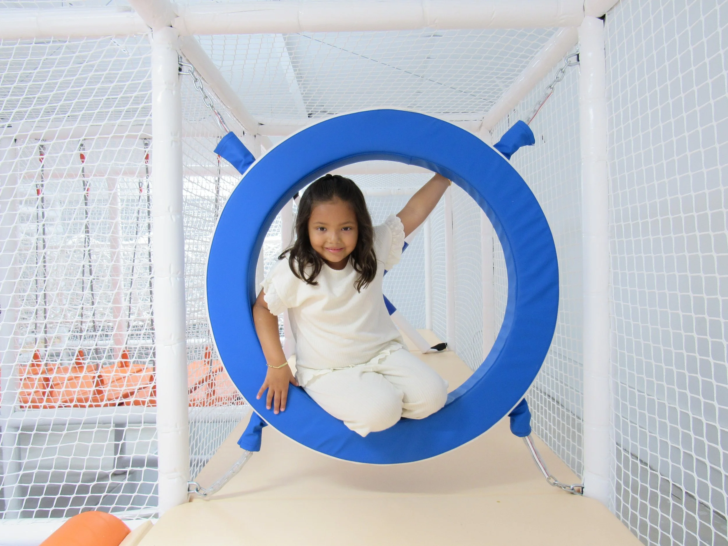 A young girl with long dark hair sitting inside a blue circular tunnel on a white play structure surrounded by white netting, smiling at the camera.