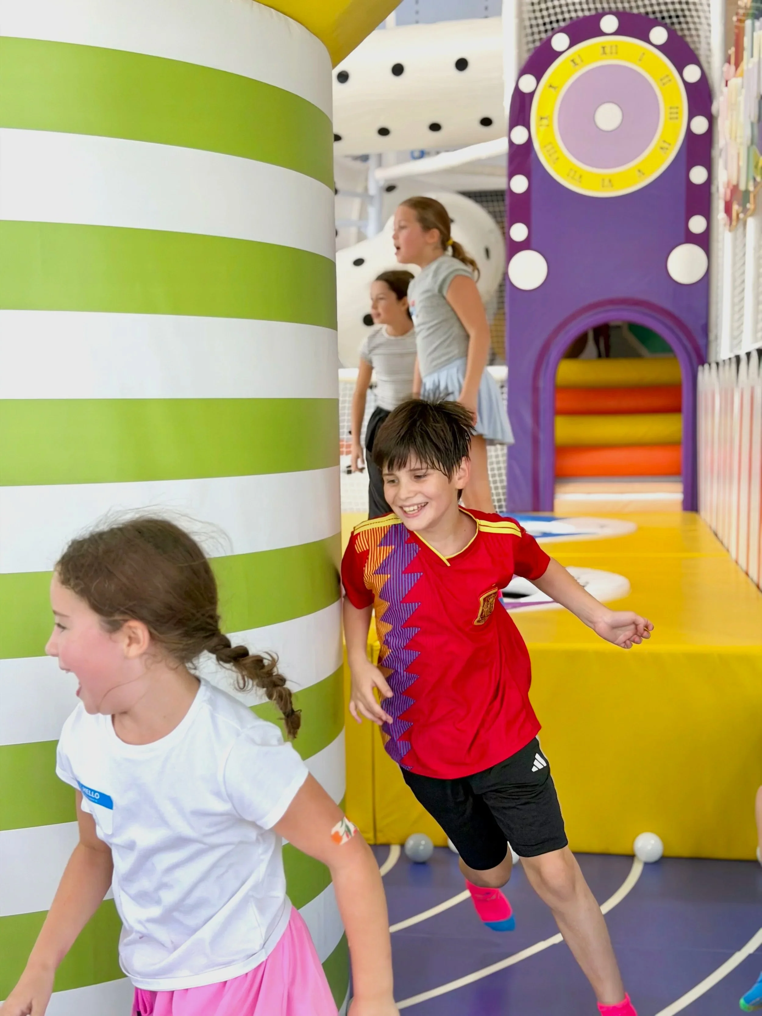 Children playing at an indoor playground with colorful equipment, including a green and white striped pillar and purple structure with yellow and orange rollers.