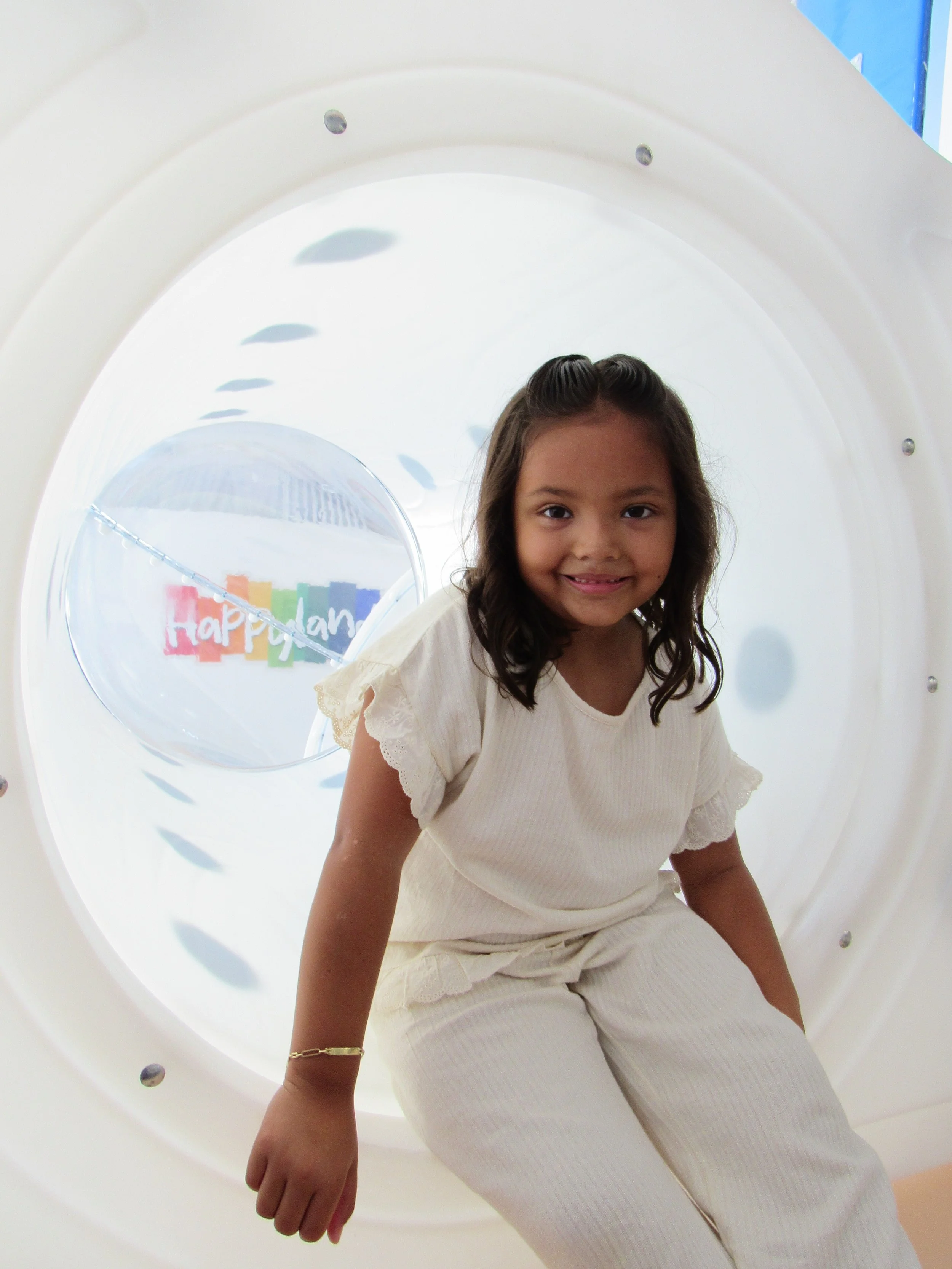 A young girl with dark hair, wearing a cream-colored outfit, posing in front of a large circular structure with a colorful 