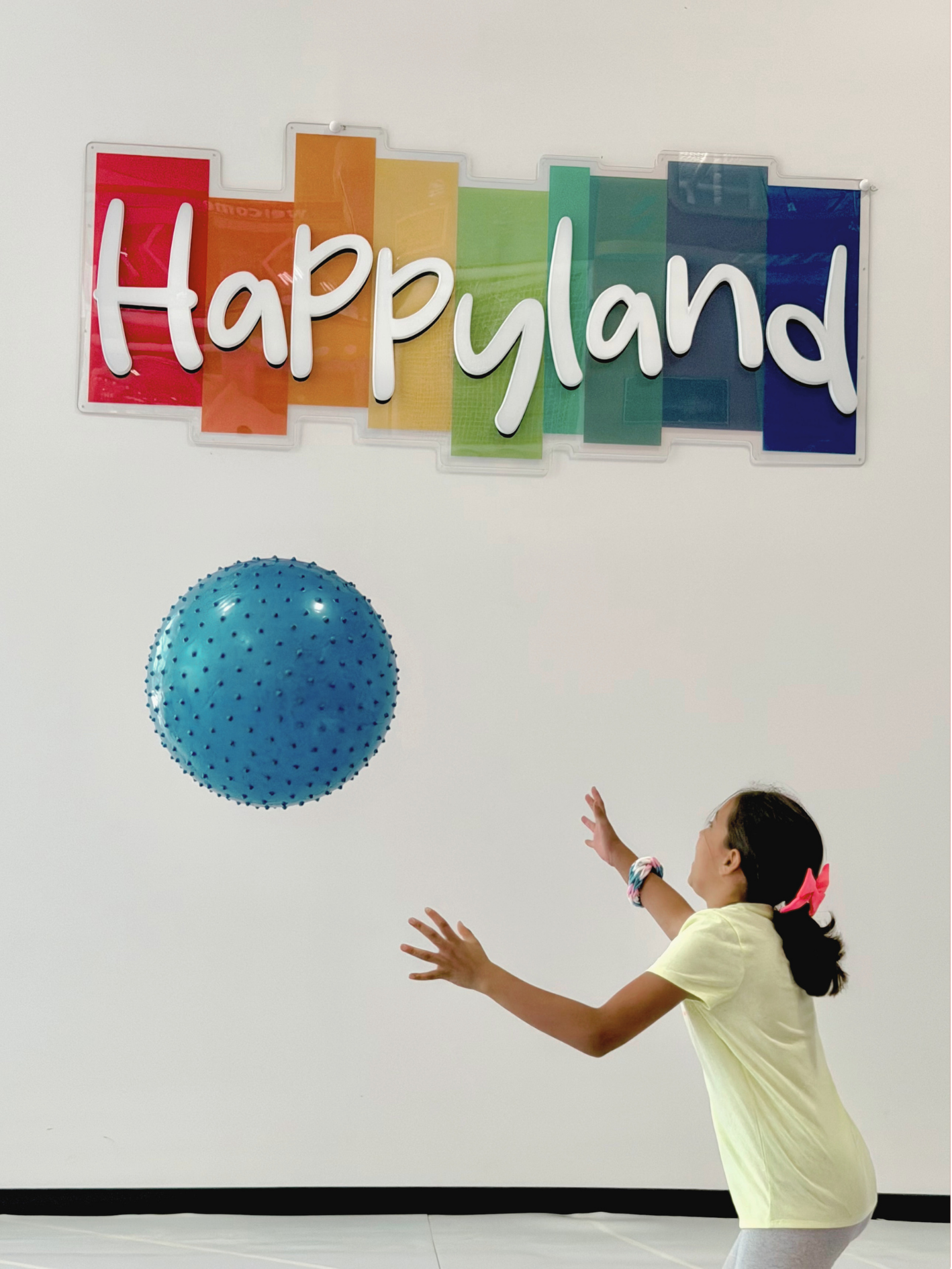 A young girl reaching out to catch or hit a large blue spiked balloon during a celebration with a colorful rainbow sign that reads "Happy Land" on a plain white background.