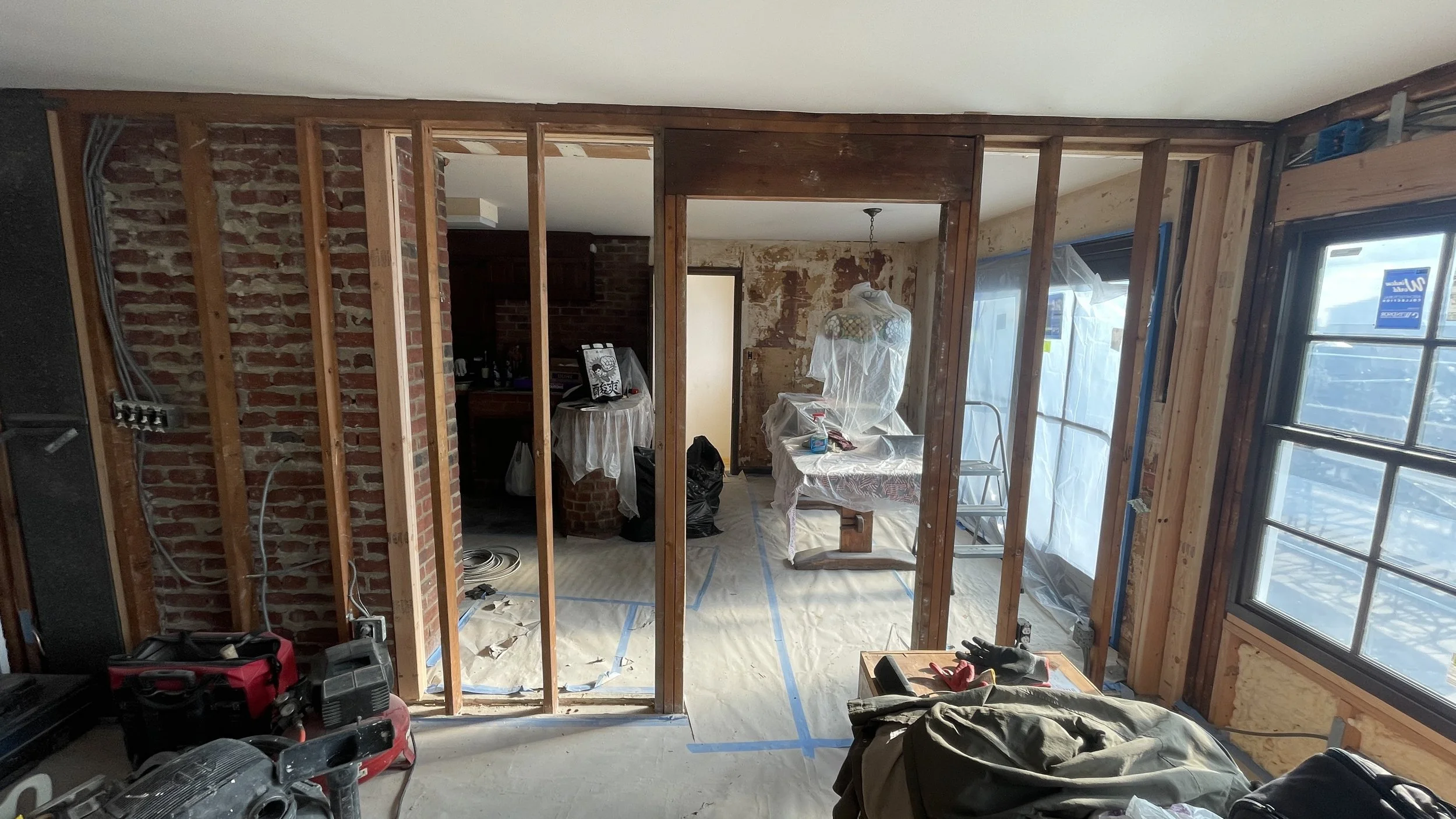 Interior of a room under renovation with exposed wooden framing, brick walls, a window, and construction tools and materials.
