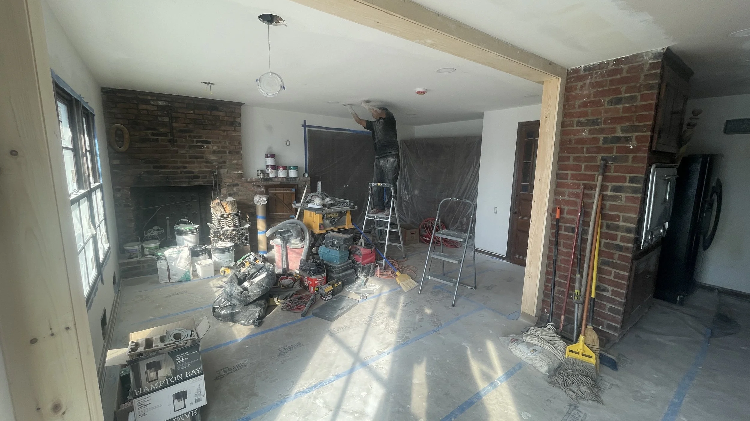 Interior of a room under renovation with tools, paint cans, and construction materials scattered on the floor, a worker on a ladder working on the ceiling, and exposed brick walls.