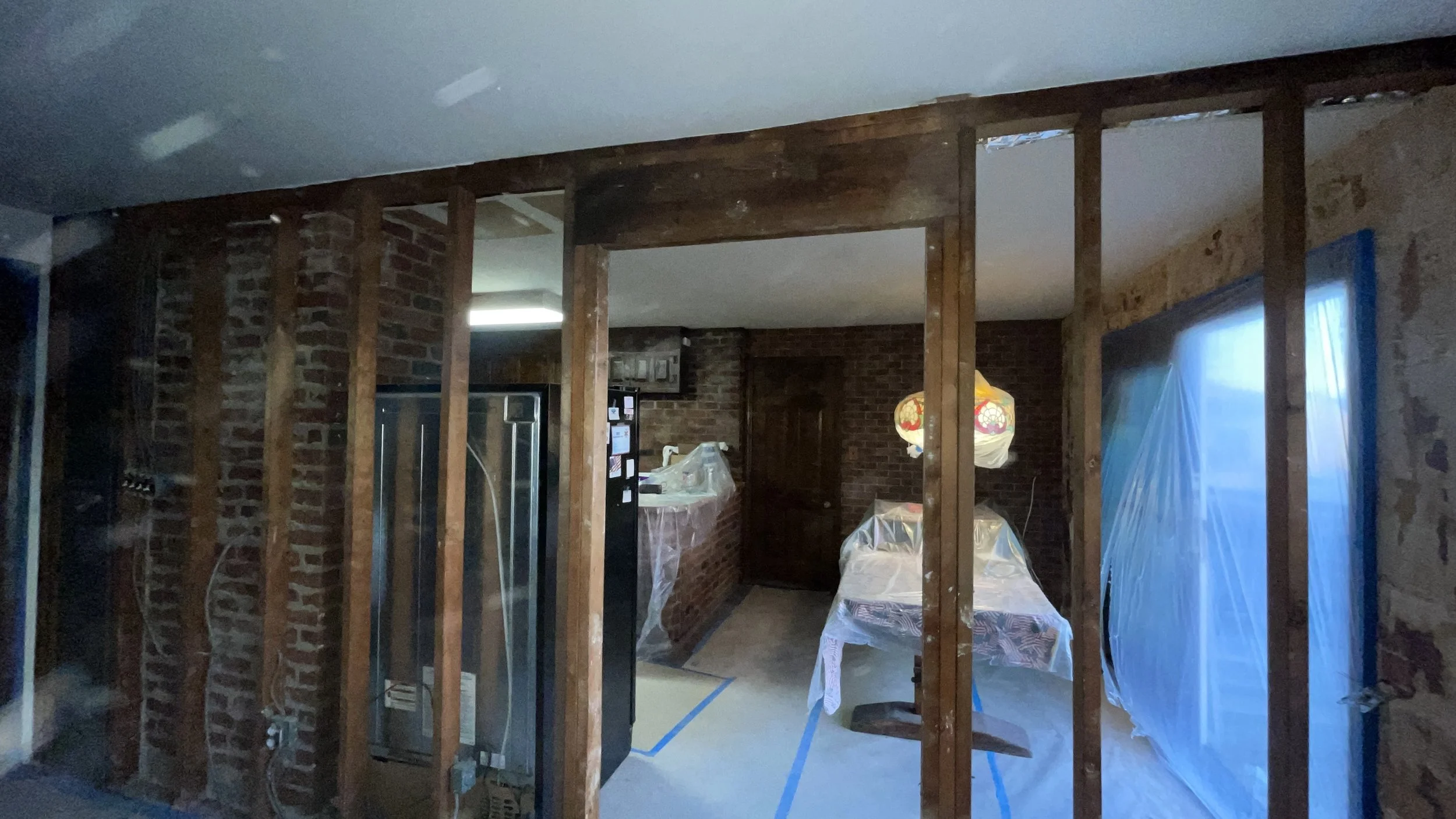 Living room undergoing renovation with exposed wooden studs, brick walls, and covered furniture, including a table with a tablecloth and a hanging light fixture.