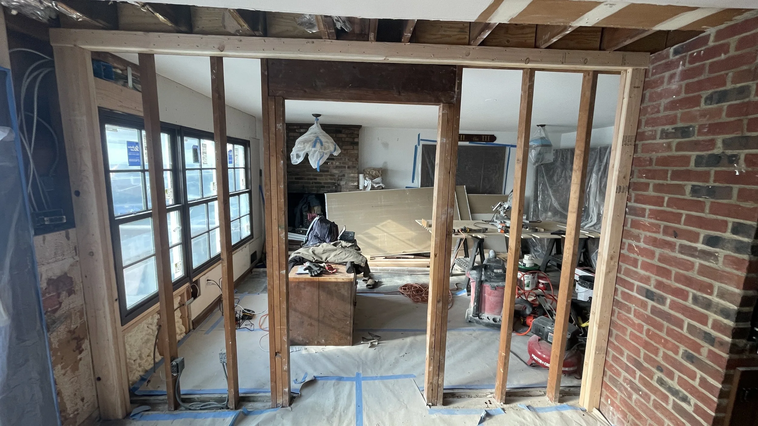 Interior of a house under renovation with exposed wooden framing, construction tools, and work materials, including a brick wall on the right and window on the left.