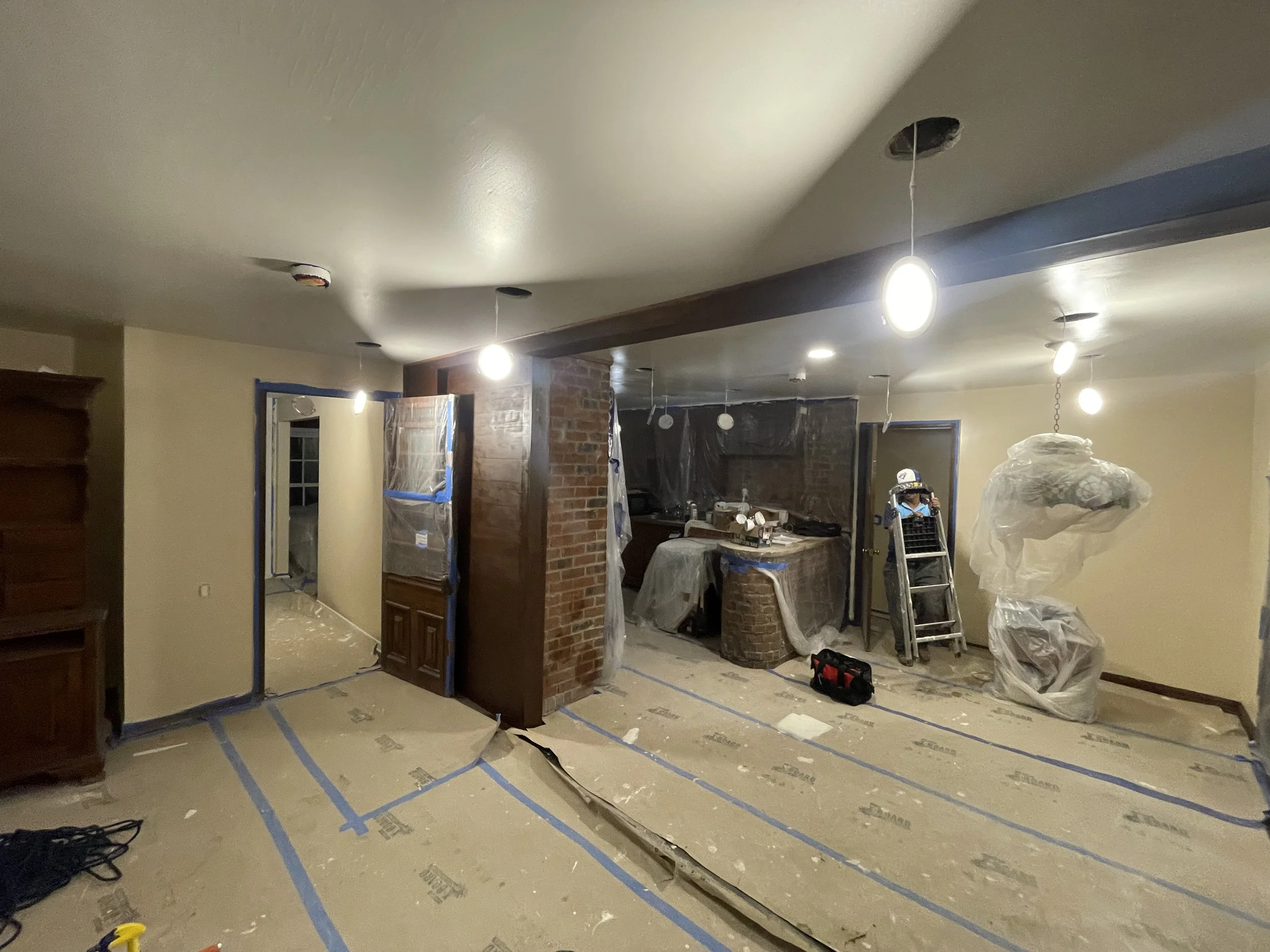 Living room undergoing renovation with taped floors, plastic-wrapped furniture, ceiling holes with hanging wires, and construction tools scattered.