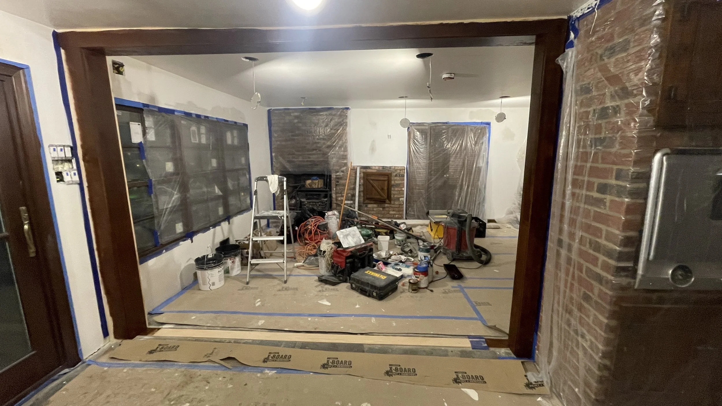 Living room under renovation with tools, construction materials, plastic sheets, and partially painted walls.