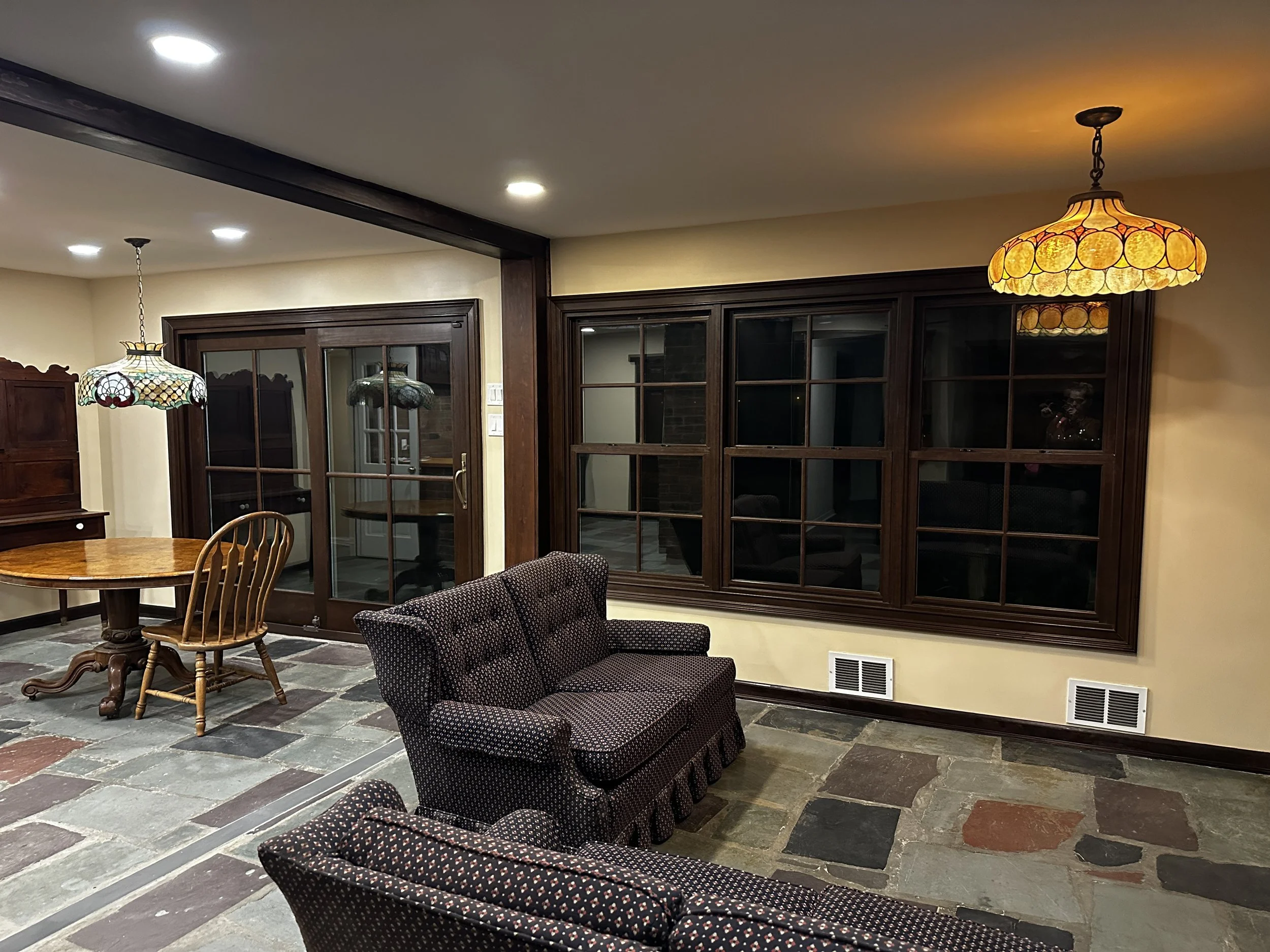Interior of a room with dark wooden framed glass doors, a multicolored tiled floor, and seating including a patterned armchair and chairs around a wooden table. Two stained glass pendant lights hang from the ceiling, illuminating the space.