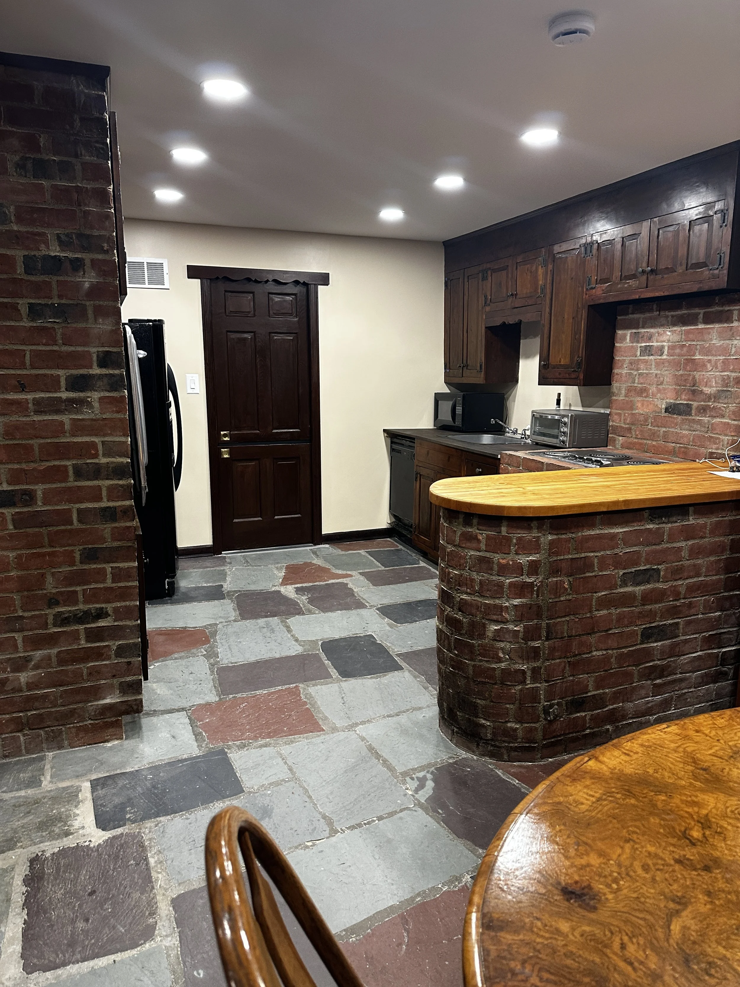 A kitchen with brick walls, wooden cabinets, a door, and a stone floor. There is a round wooden table and chairs in the foreground. Appliances such as a microwave and toaster are on the counter.
