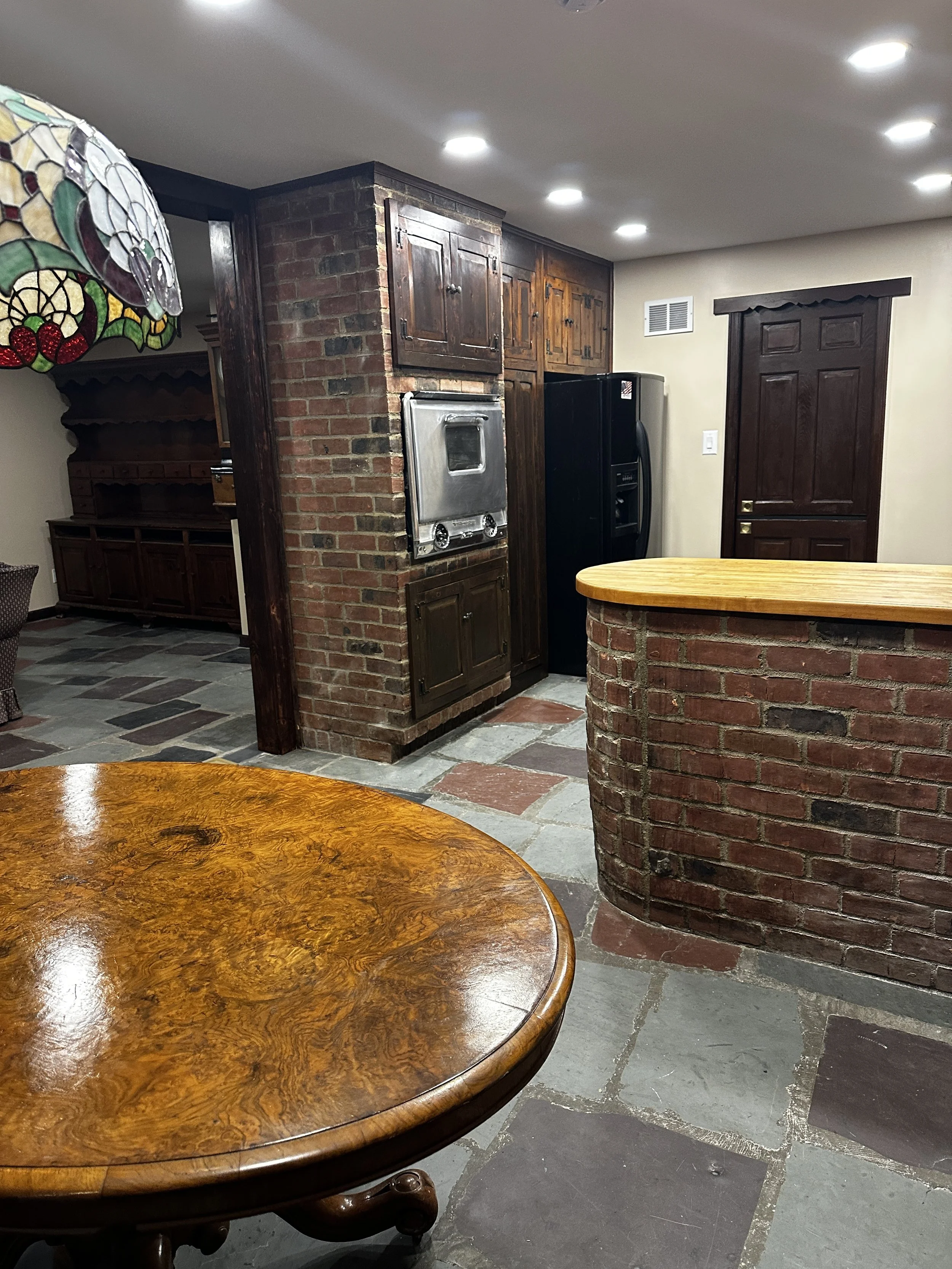 Interior of a cozy room with a brick and wood bar counter, a vintage wooden cabinet, a black refrigerator, and a table with a polished wooden surface. Ceiling lights illuminate the space, and there is a decorative stained glass lamp partially visible