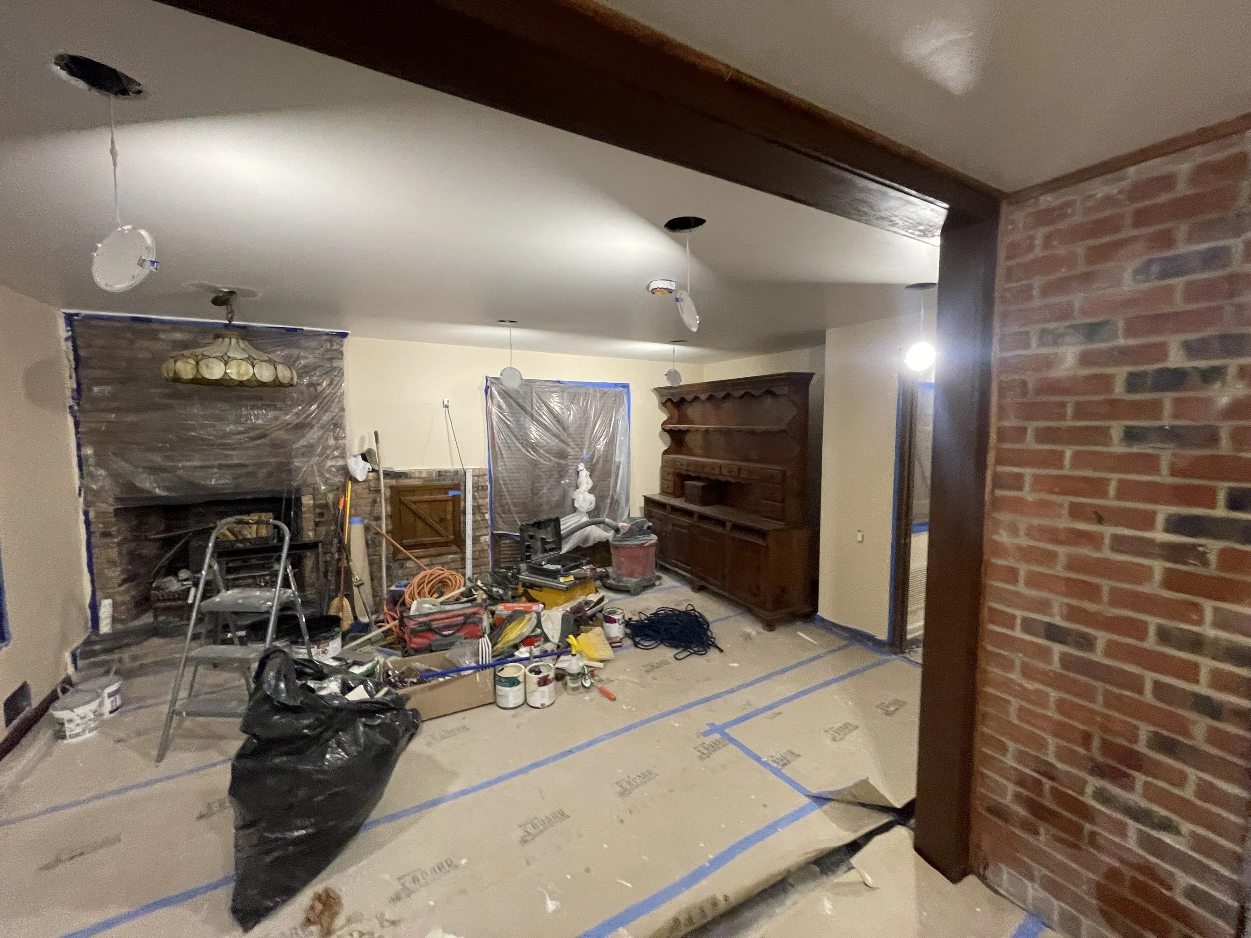 Living room under renovation with painting supplies, tools, and protective plastic covering on the floor. Exposed ceiling with light fixtures, brick wall, and partially finished area.