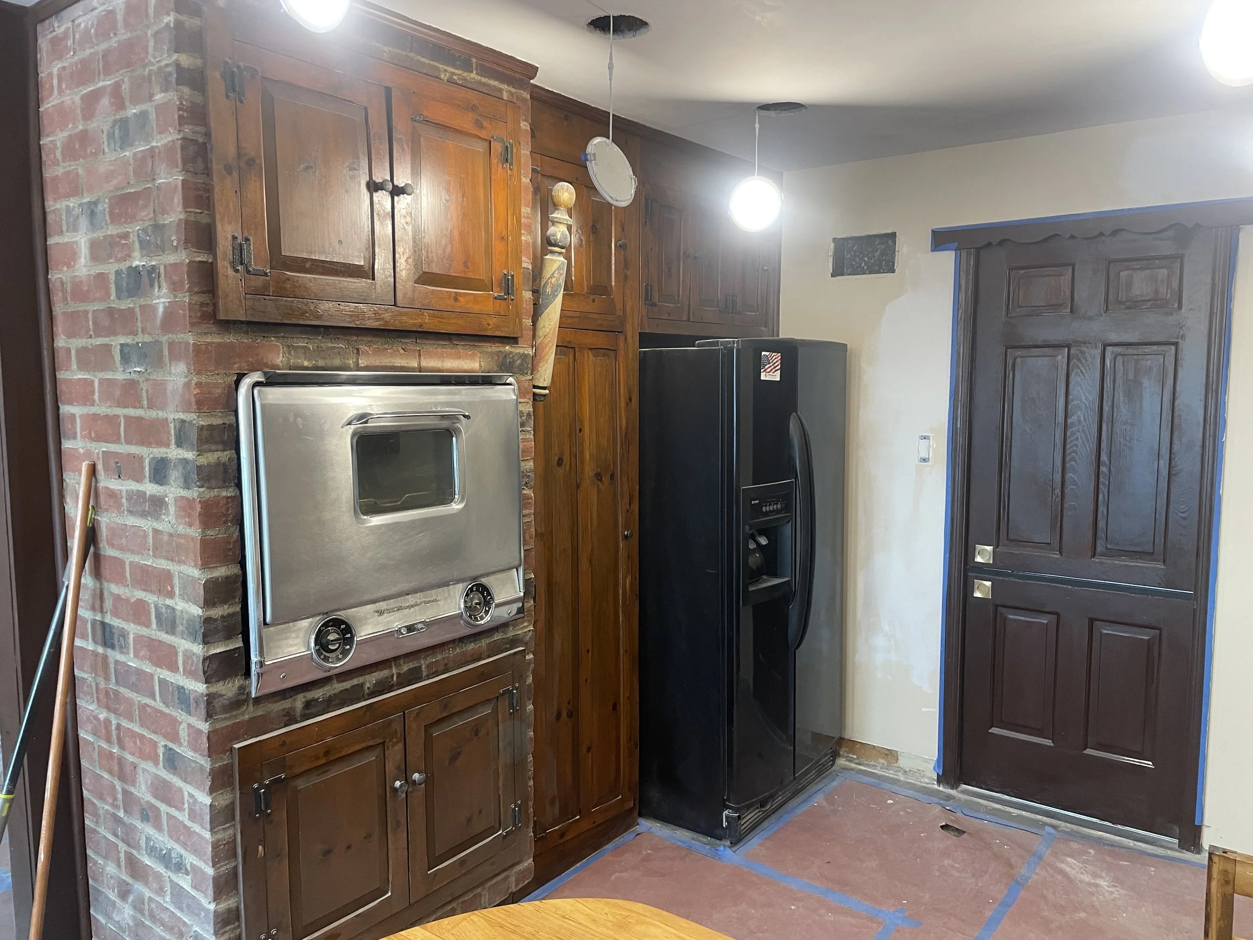 A kitchen wall with brick fireplace, wooden cabinets, black refrigerator, and a stainless steel oven with a glass window. The floor has blue tape outlining an area, and part of a dark wooden door is visible on the right.