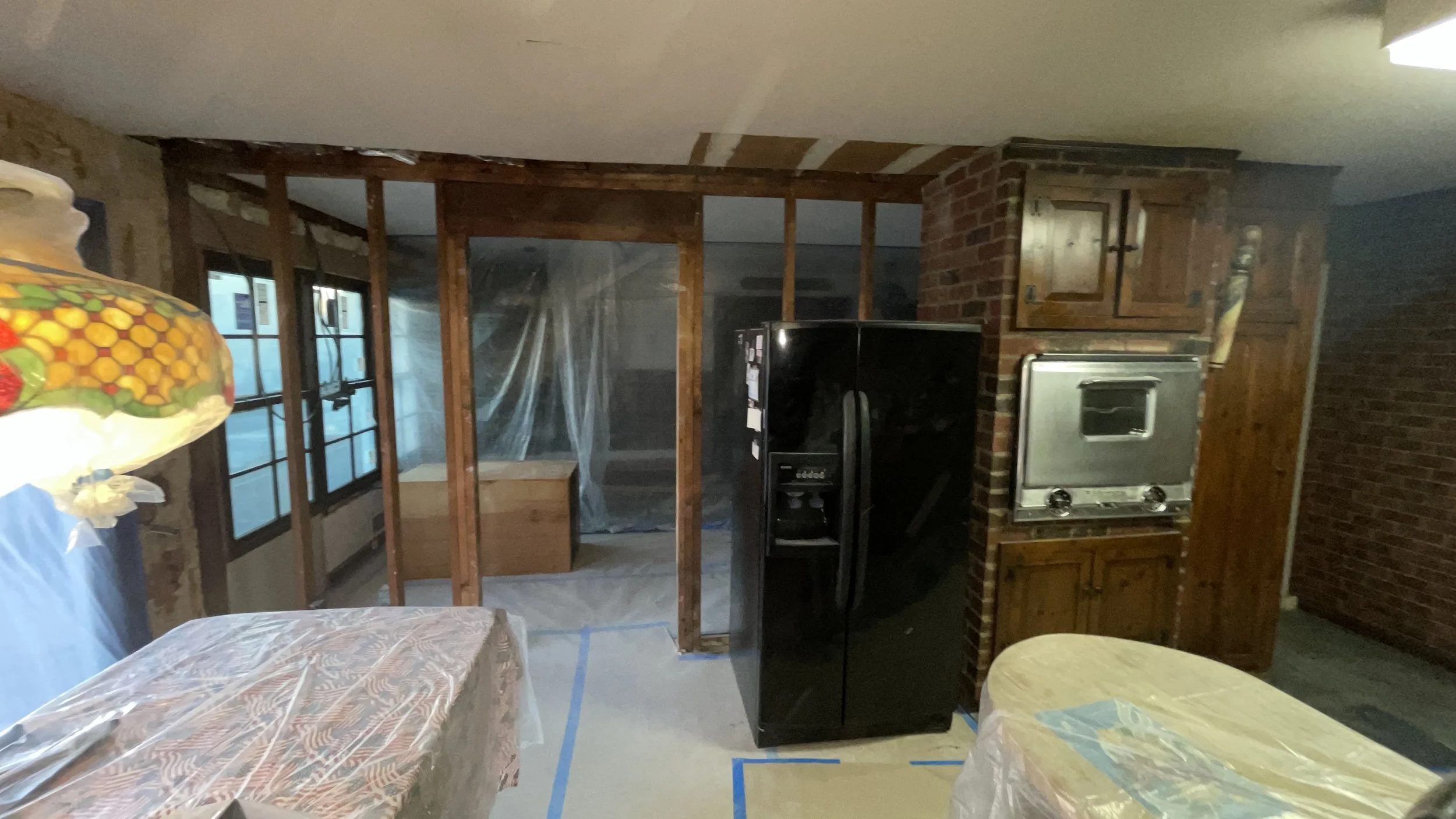 Partially constructed kitchen with exposed wooden framing, brick and wood cabinets, black refrigerator, and a double oven, with plastic-covered tables and blue painter's tape on the floor.