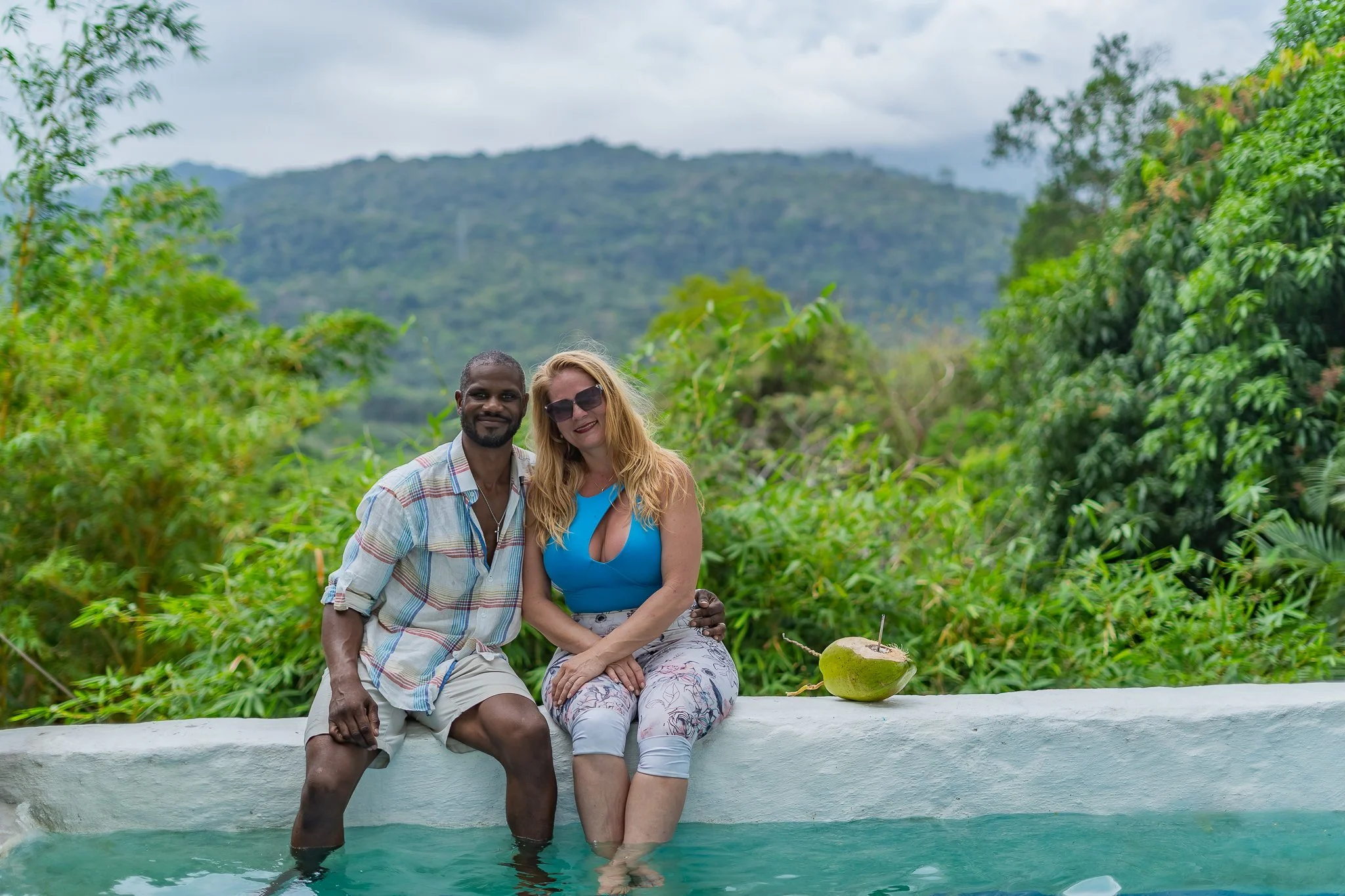 A couple sitting by a pool with a lush green mountain background. The man has dark skin, short hair, and is wearing a plaid shirt with beige shorts. The woman has fair skin, long blonde hair, and is wearing sunglasses and a blue top. A coconut is on the ledge beside them.