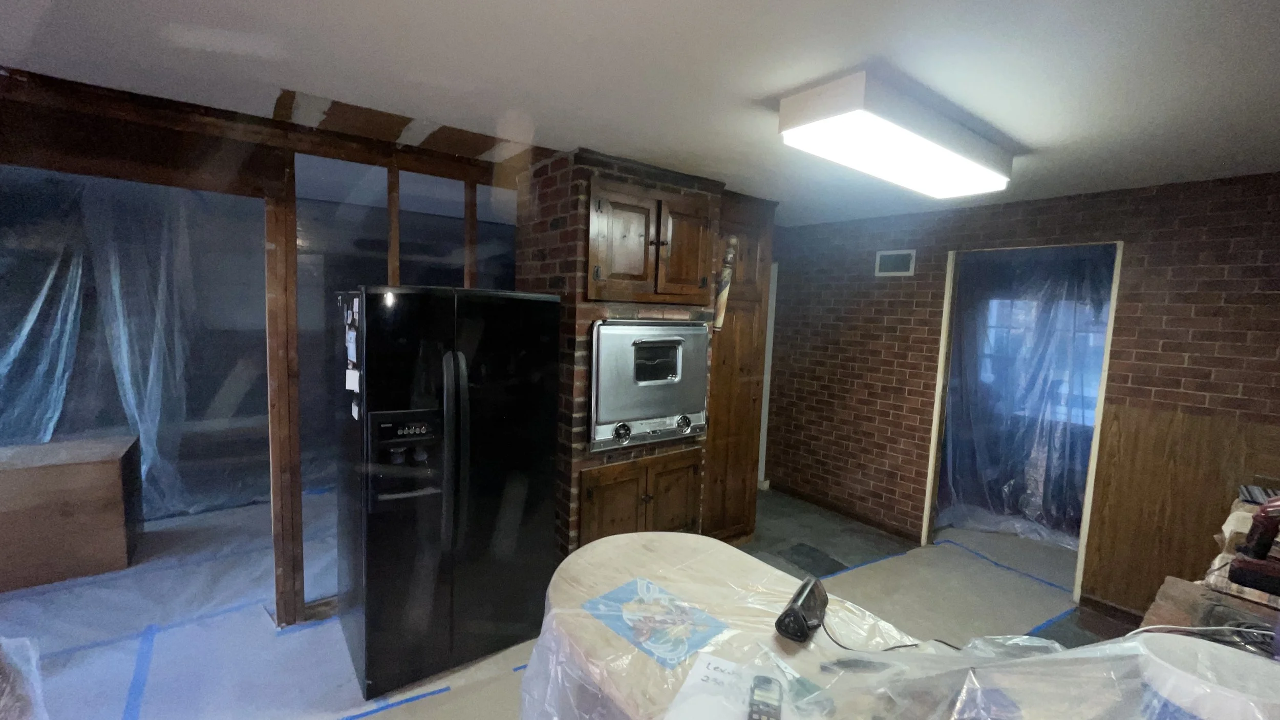 Interior of a room under construction with exposed brick walls, wooden cabinets, a black refrigerator, and a stainless steel oven. Large windows covered with plastic, blue painter's tape on ceilings and floor, and a round table with a cloth cover and