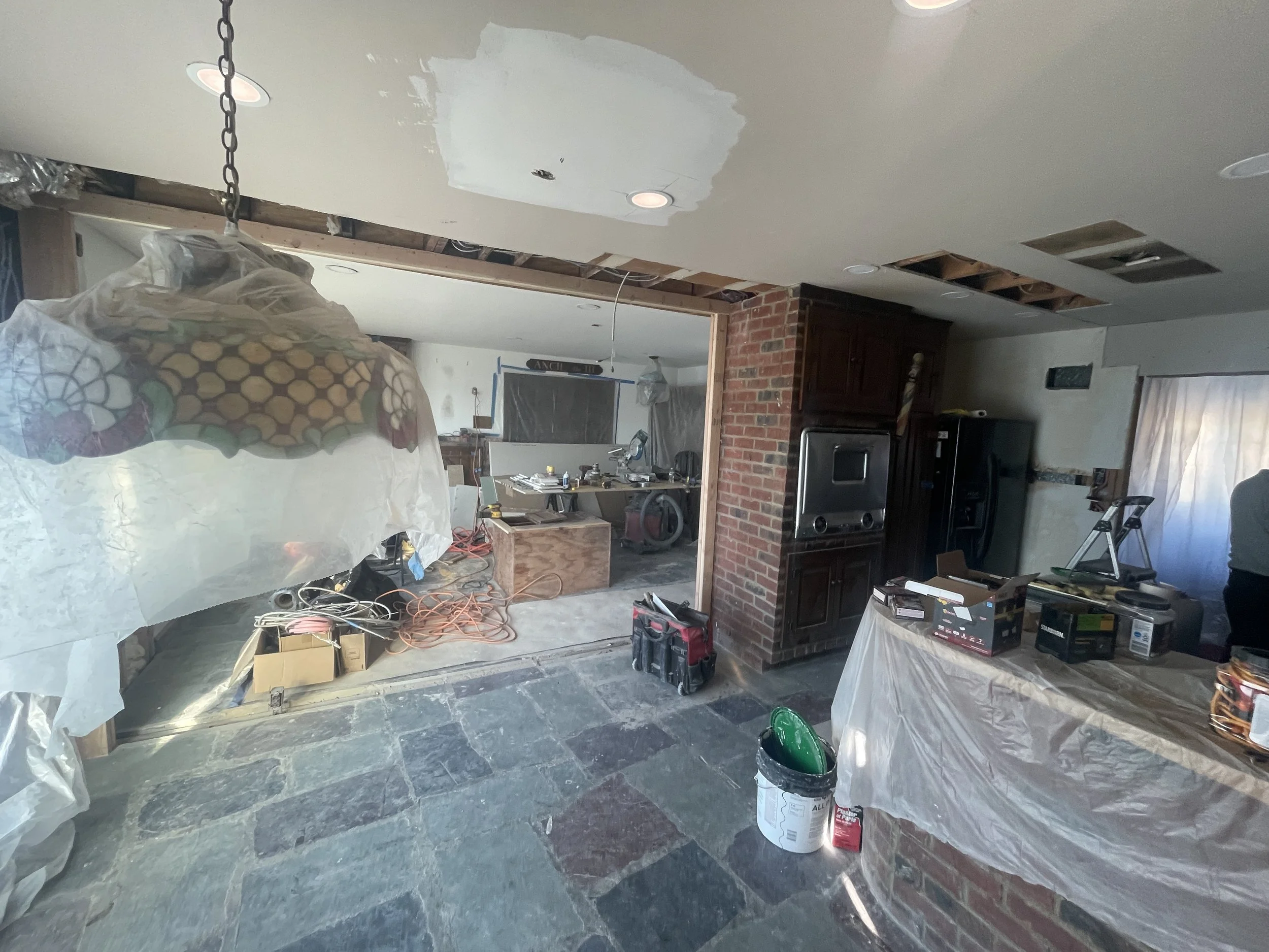 Living room undergoing renovations with drywall installation, construction tools, and supplies visible, including plastic-covered furniture, electrical cords, and a brick fireplace.