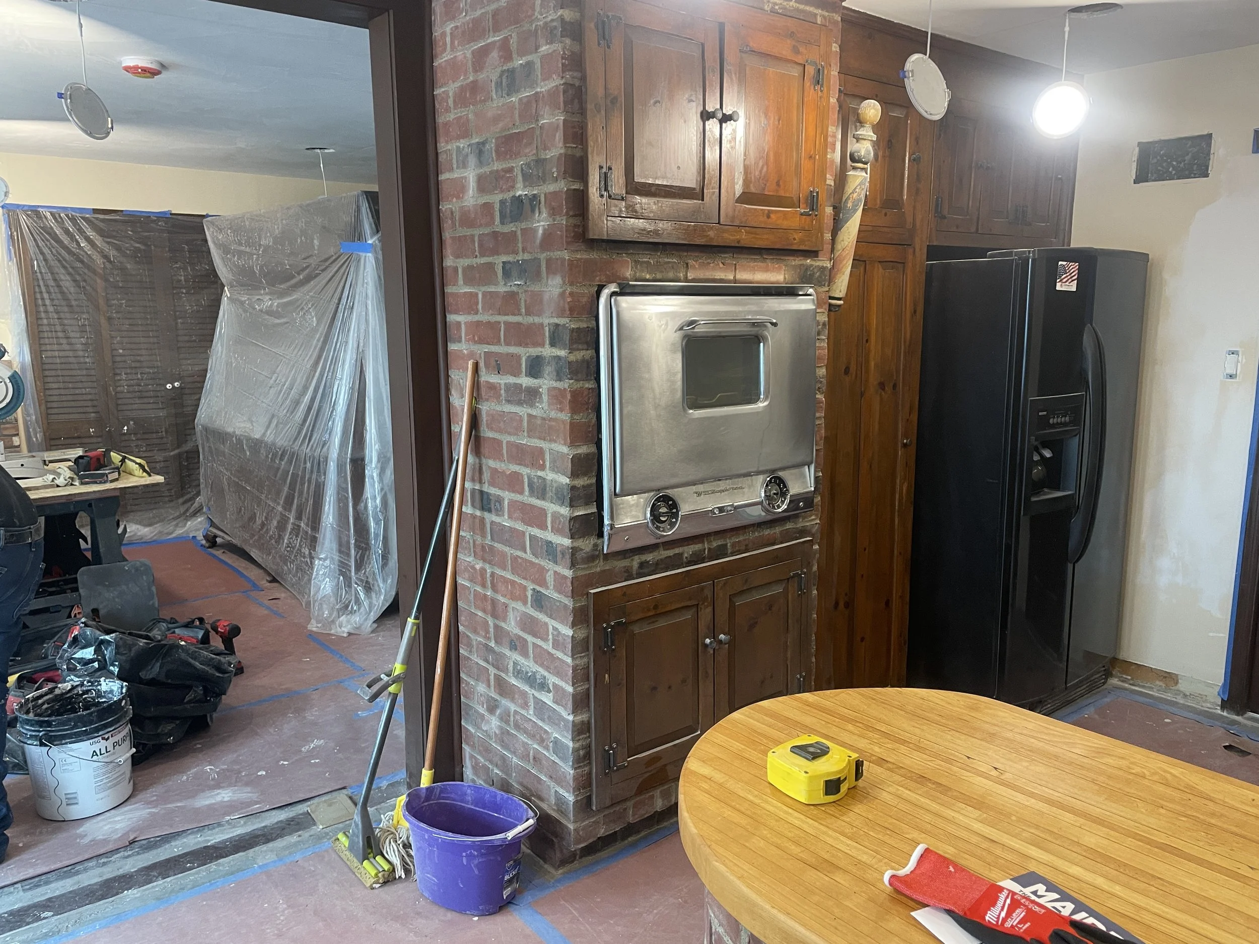 Kitchen with unfinished renovation, tools, brown cabinets, black refrigerator, brick wall, plastic-covered furniture, construction supplies, and a round wooden table.